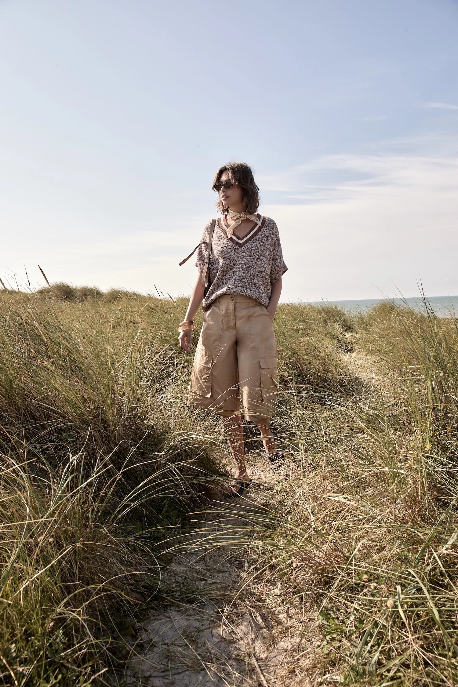 Woman walking through tall grass dunes near the beach, wearing sunglasses, a patterned sweater, beige pants, and carrying a shoulder bag.