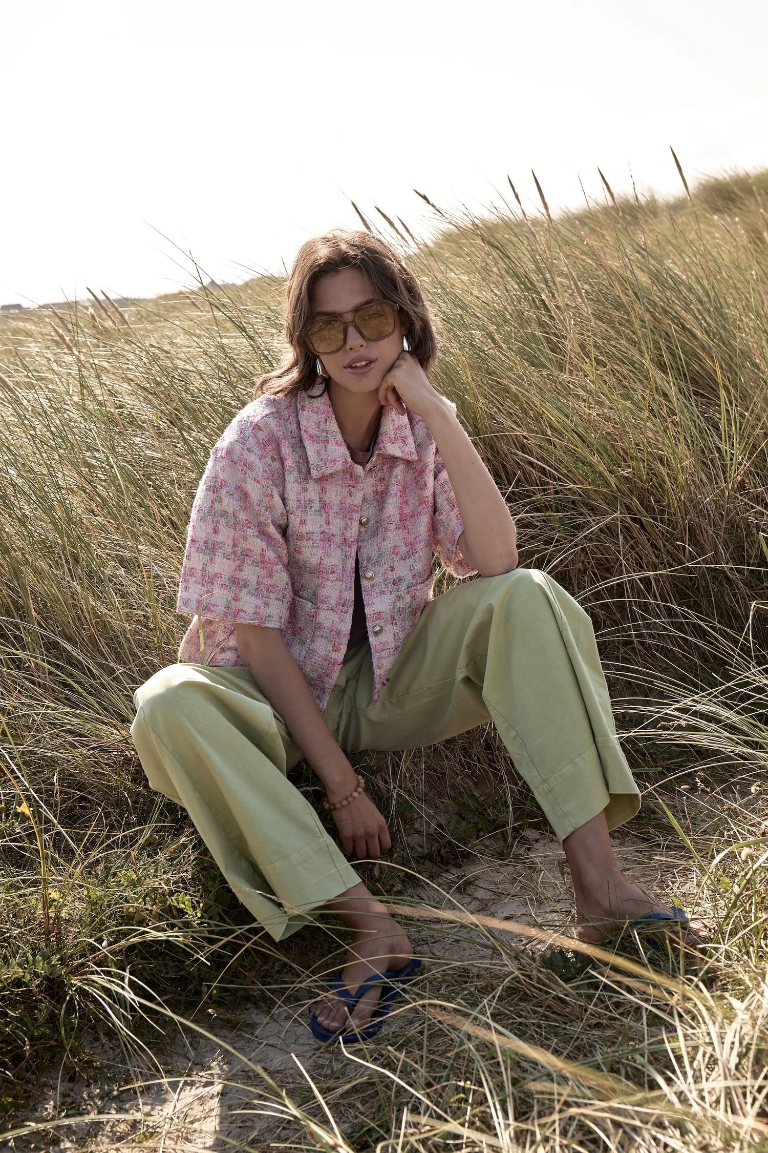 Woman sitting in tall grass wearing sunglasses, pink patterned shirt, and beige pants, with a hand resting on her chin.