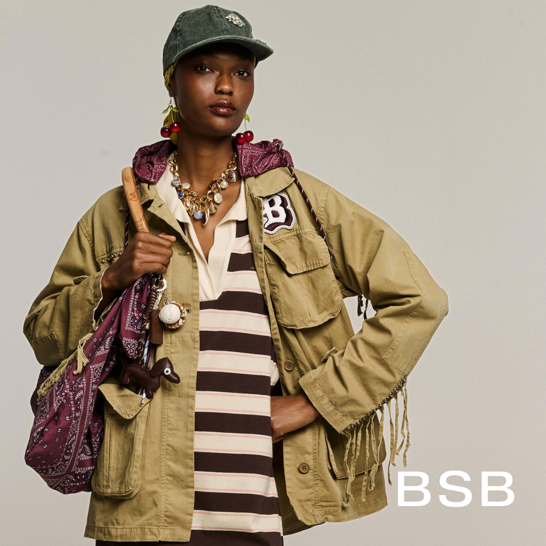 A woman with dark skin wearing a green cap, earrings with cherries, and layered necklaces. She has on a beige jacket with a patch on the chest, over a striped shirt. She carries a bag with bandanas, and a keychain with a small dog figure. The background is plain and neutral.