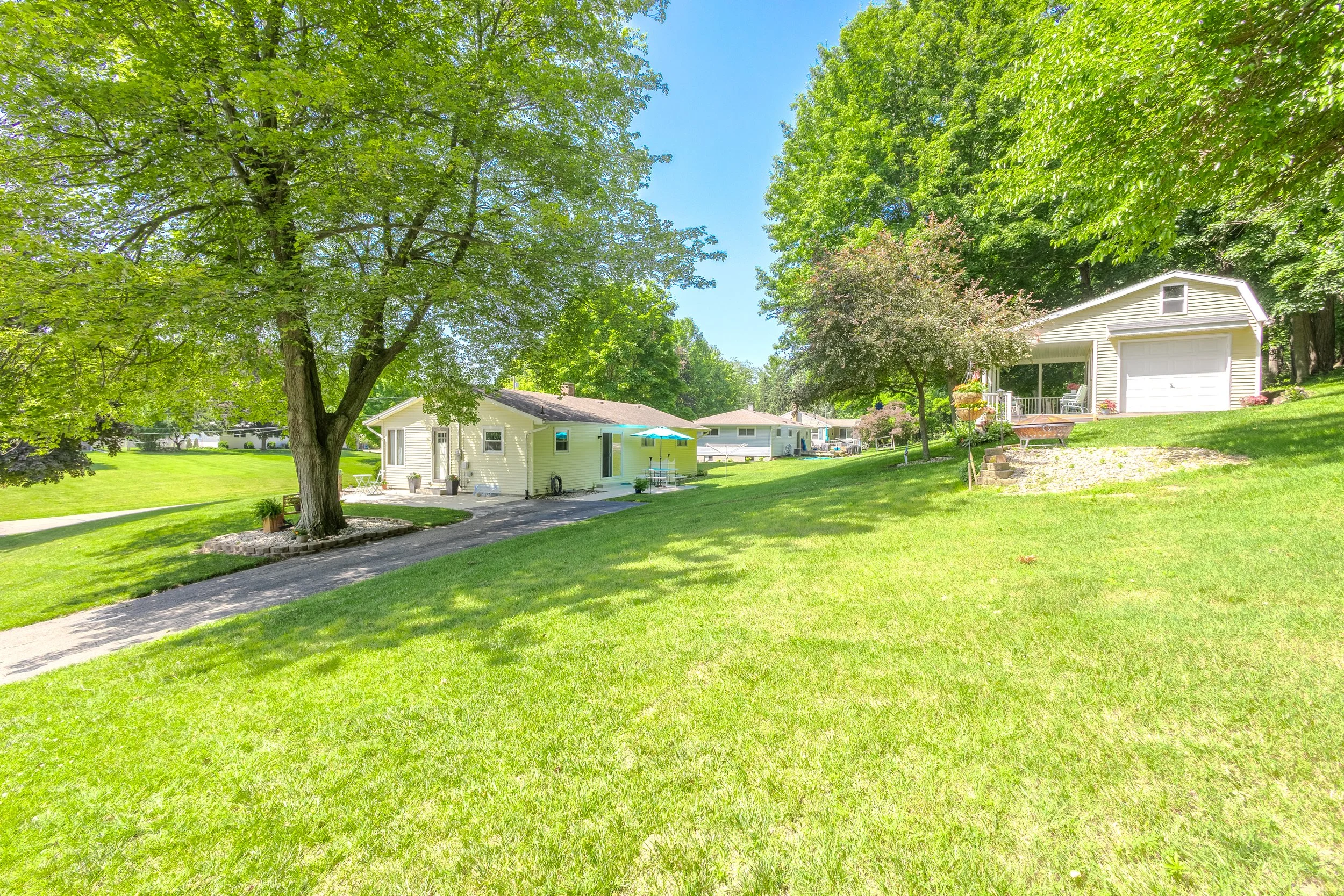 A lush green backyard with a large tree, a small house, a detached garage, and a stone-lined planting bed on a sunny day.