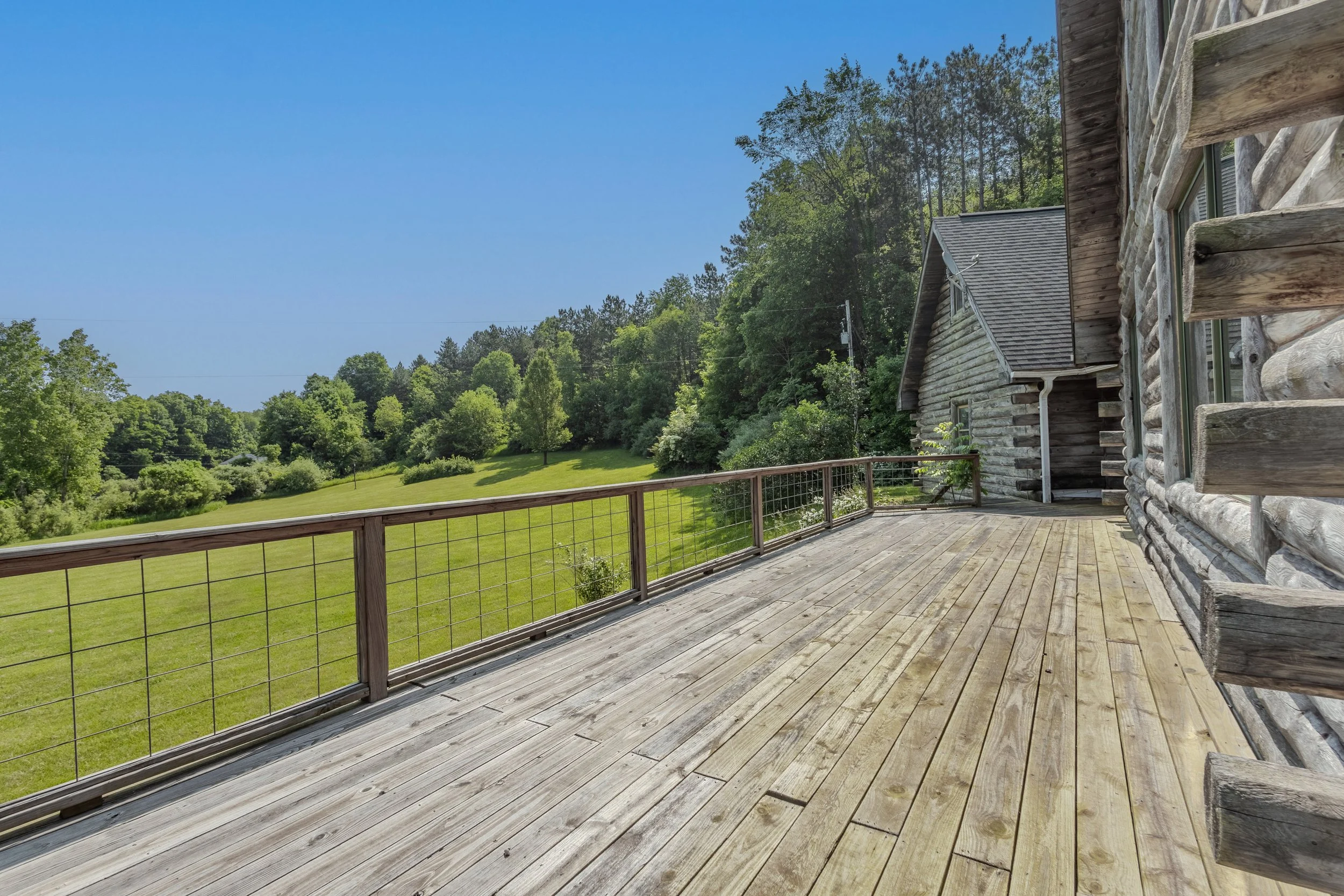 Wooden deck attached to a log cabin with a view of a green lawn and trees in the background under a clear blue sky.