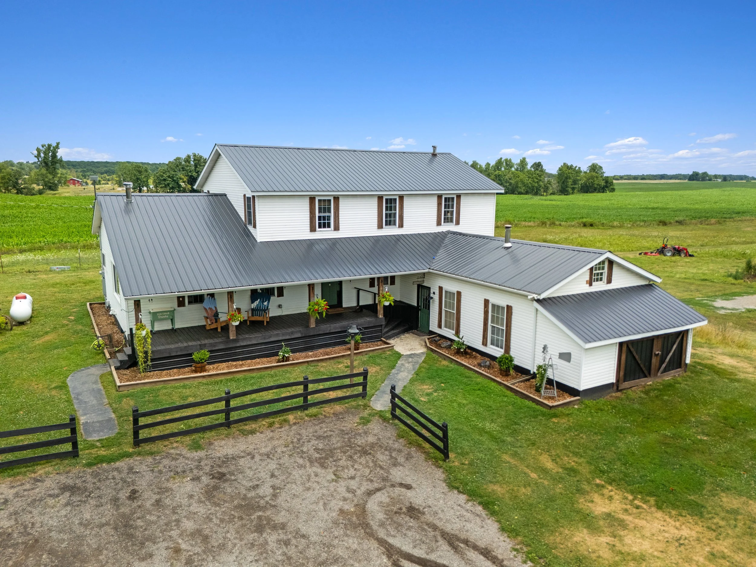 A large white farmhouse with multiple sections and gray metal roofs, surrounded by green fields and a partly cloudy sky.