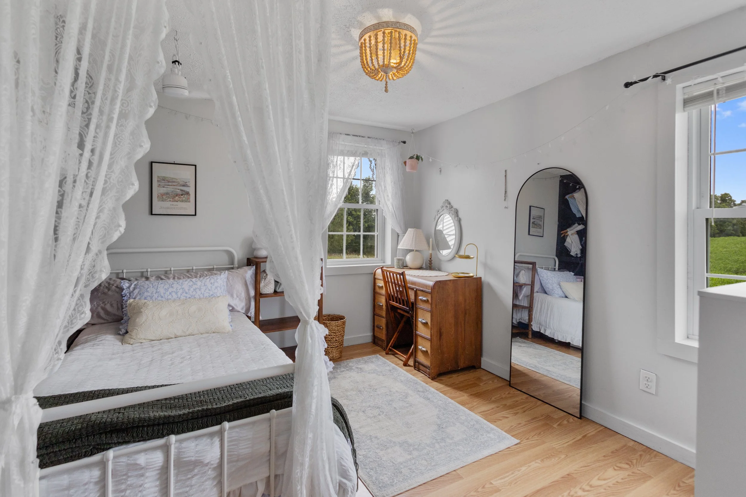 A cozy bedroom with a white four-poster bed, lace curtains, a wooden desk with a mirror, a tall mirror, a window with white curtains, and a wood floor.