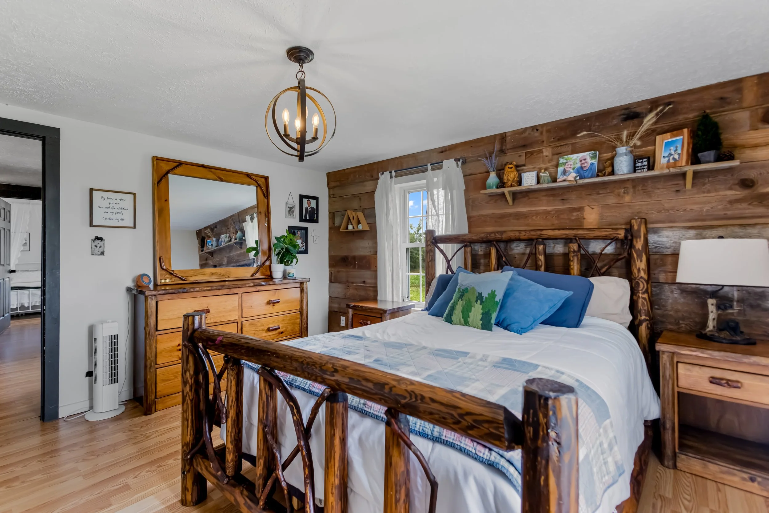 Bedroom with wooden bed frame, matching wooden dresser, and a mirror; white walls with black trim; wooden accent wall; window with white curtains; decorative shelf with photos and decor; blue pillows on bed; wooden nightstand with lamp and phone; cei