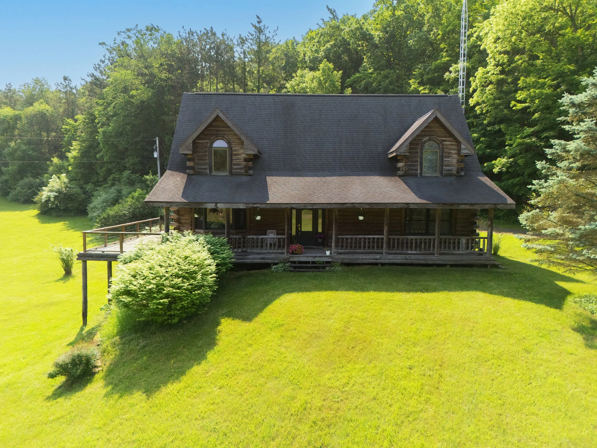 A rustic wooden house with a large front porch, surrounded by green grass and trees, under a clear blue sky.