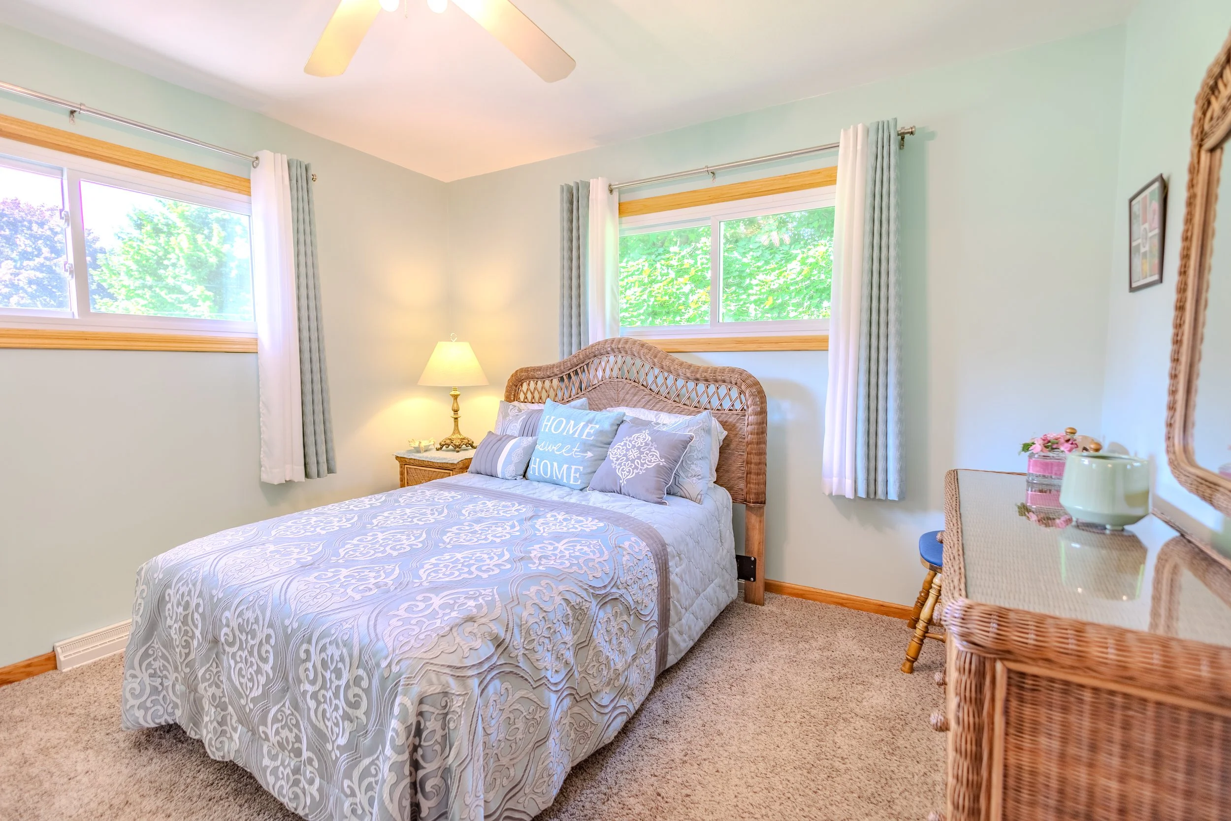A cozy bedroom with light green walls, a bed with a wicker headboard, white and blue bedding, decorative pillows, and twin windows with curtains. There is a nightstand with a lamp and a wicker dresser with mirrors.