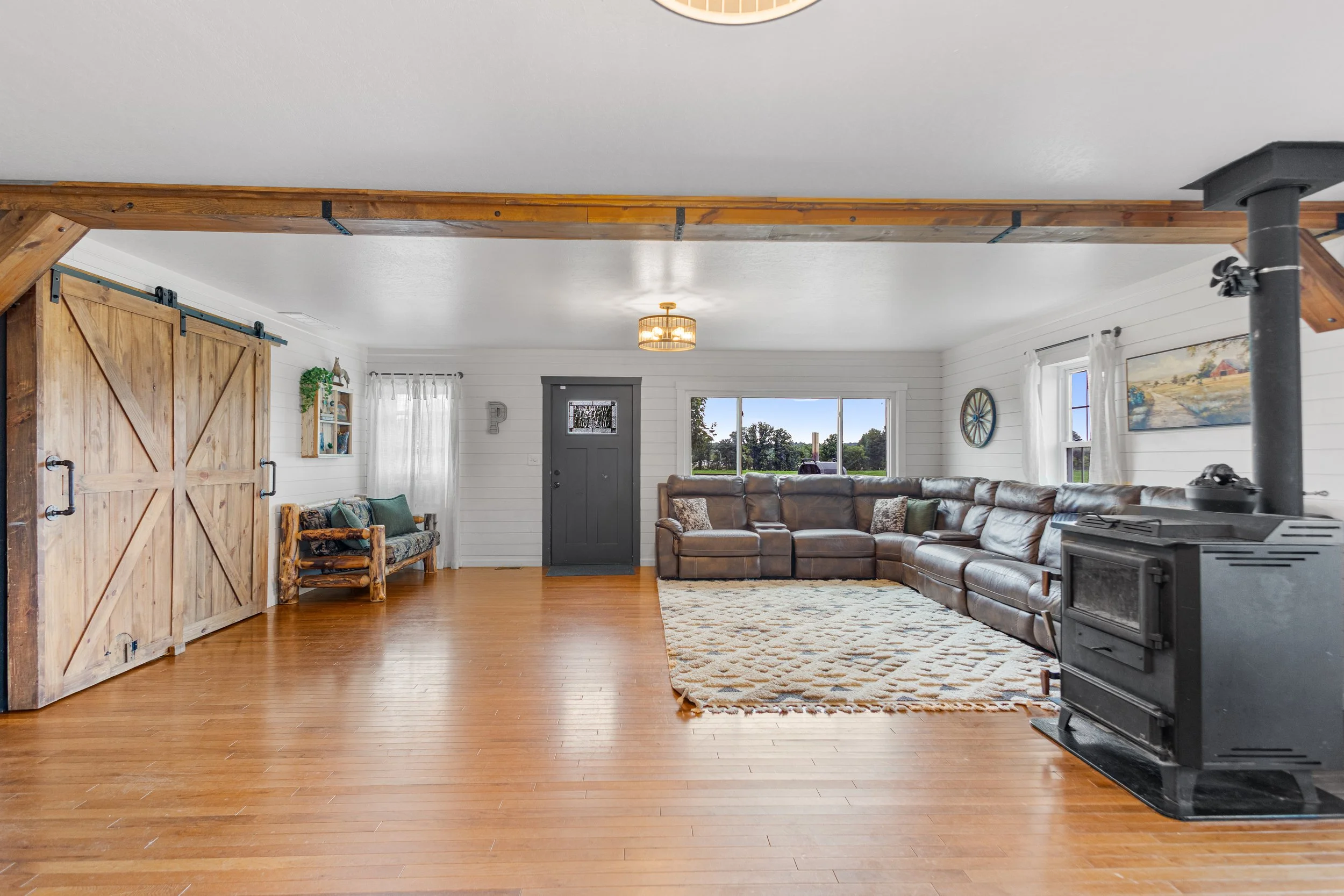 Living room with hardwood floors, white paneled walls, large brown sectional sofa, wood stove, and sliding barn doors