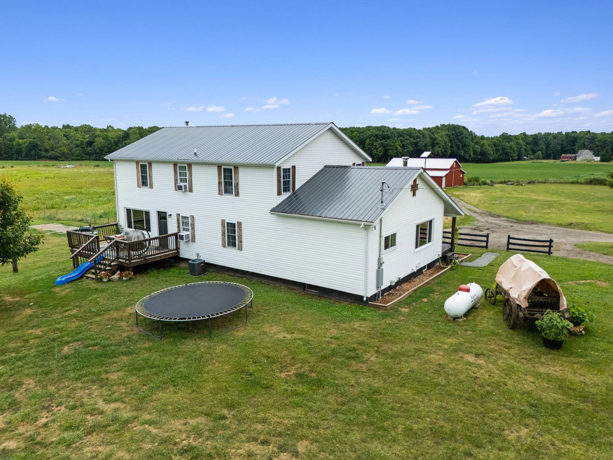 A two-story white house with a metal roof, a wooden deck with a slide, a trampoline, and outdoor equipment on a grassy lawn in a rural setting with fields and barn structures in the background.