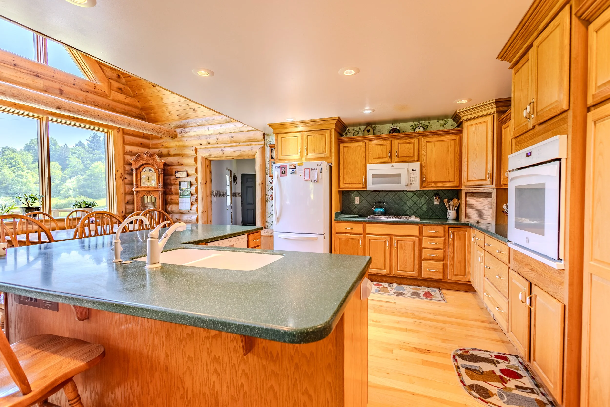 Kitchen with wooden cabinets, green countertops, white appliances, and a large window showing trees outside.