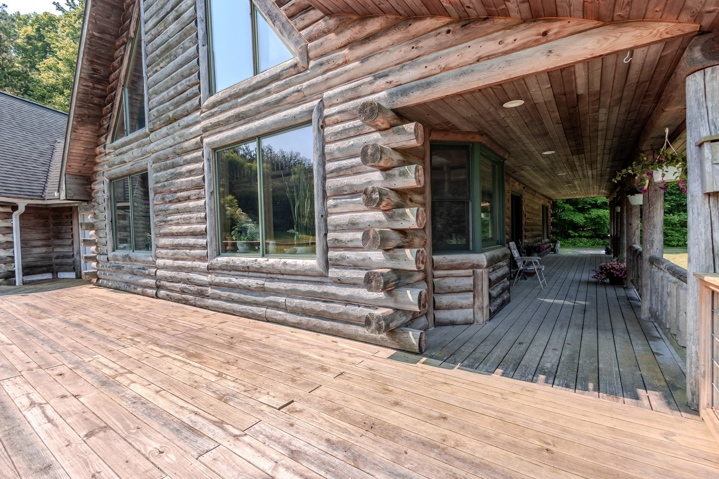 Close-up view of a rustic log cabin with a wooden deck and porch, featuring large windows and potted plants, surrounded by green trees.
