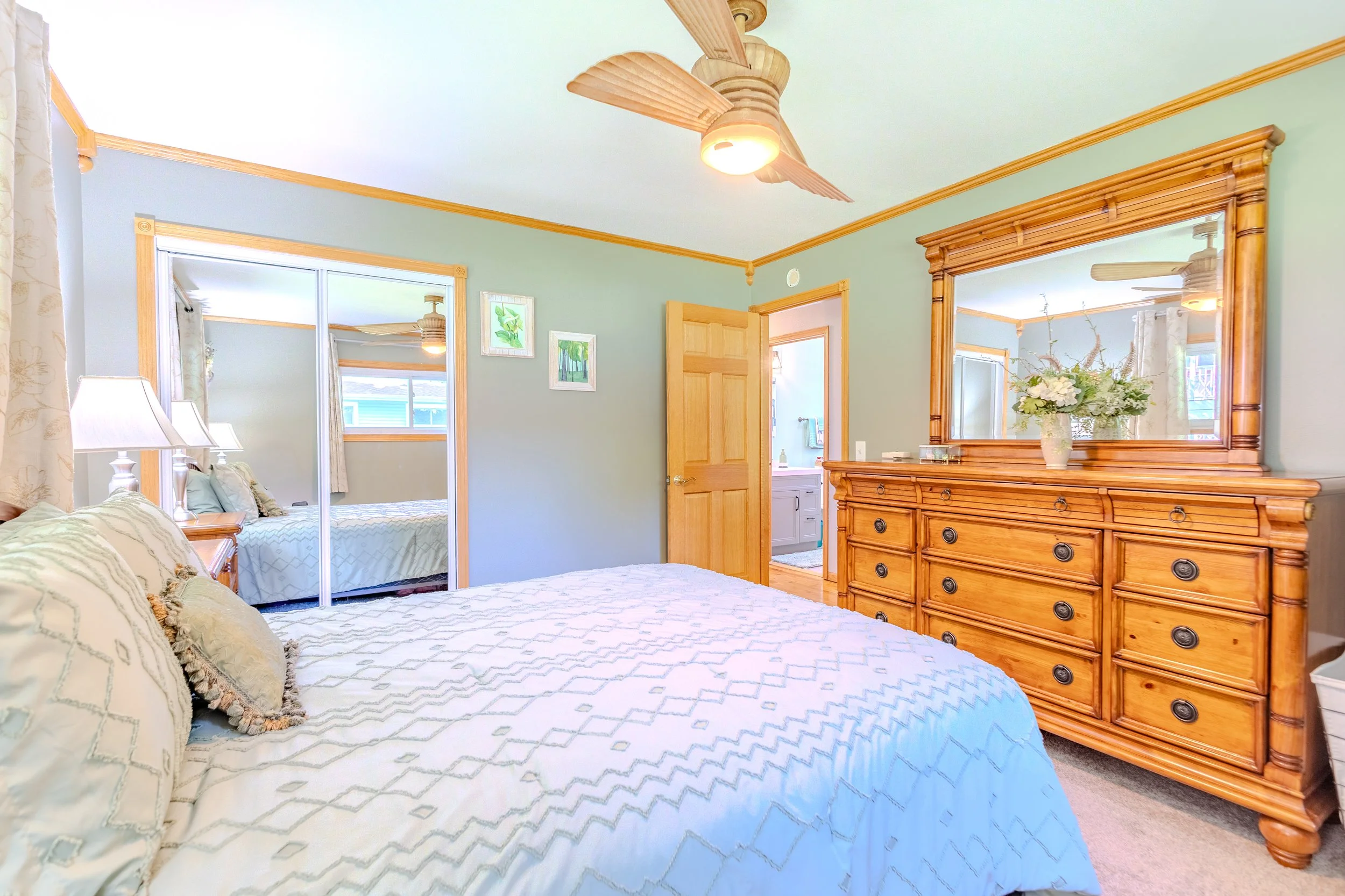 Bedroom with a bed, wooden dresser with mirror, and a ceiling fan, with an open door leading to an adjoining bathroom.