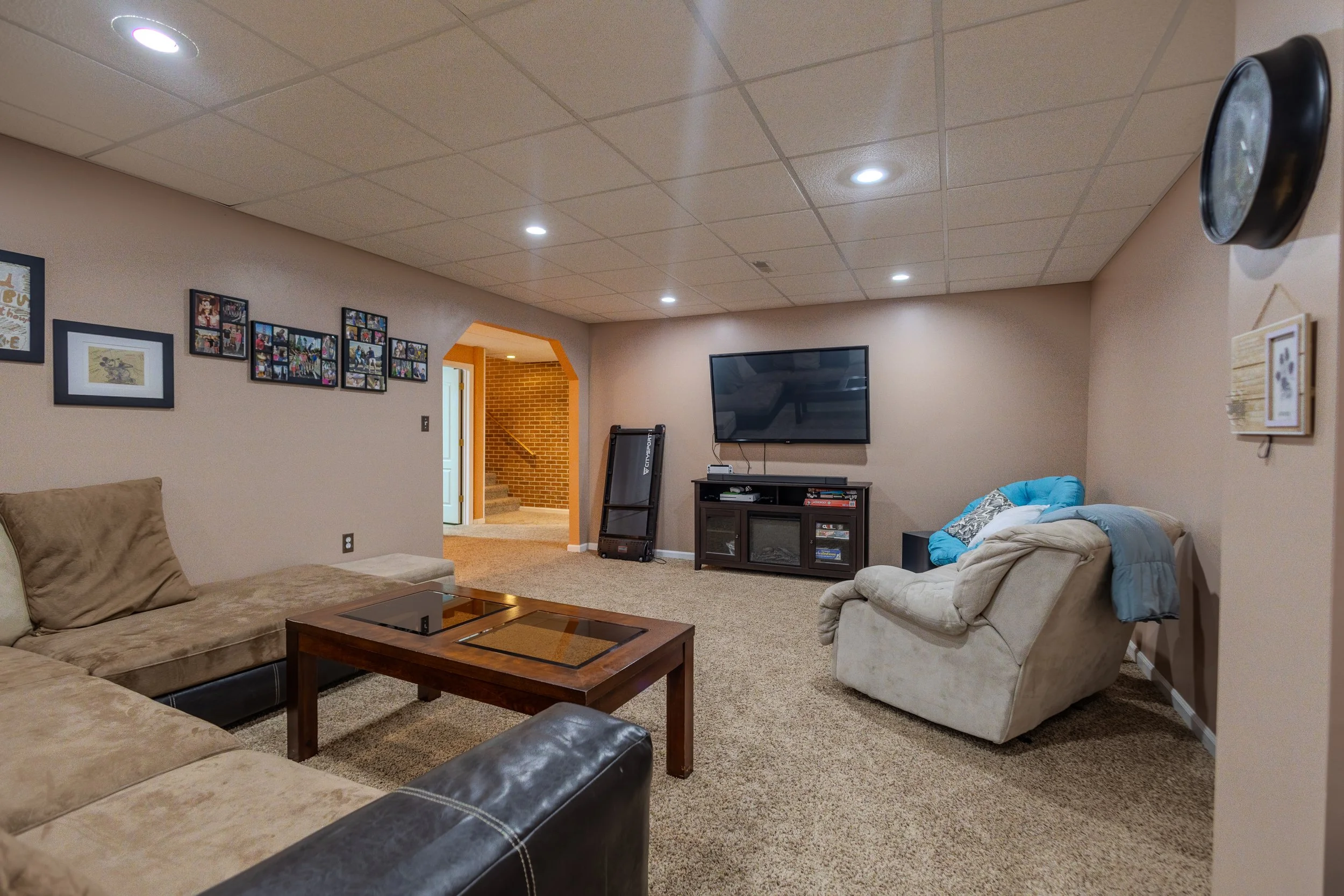 A living room with beige walls and carpet, featuring a sectional sofa, an armchair with blankets, a wooden coffee table, a mounted flat-screen TV, a media console, and framed photos on the wall. There is a staircase area visible through an open doorw