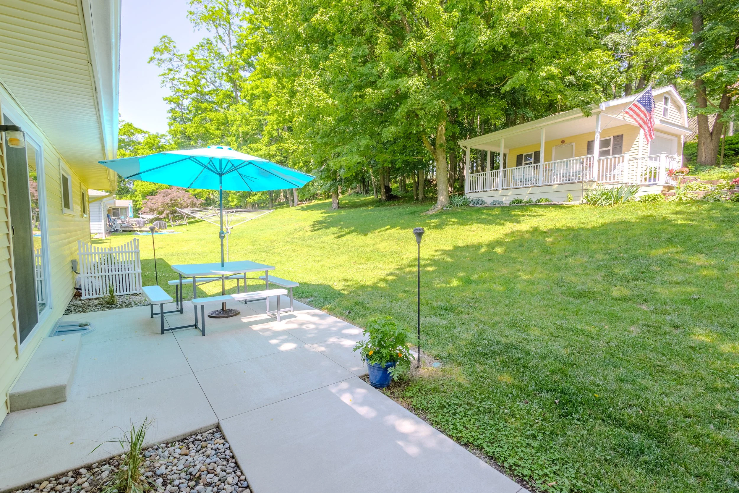 Backyard with concrete patio, blue patio umbrella, picnic table, green lawn, trees, and a small yellow house with an American flag on porch.