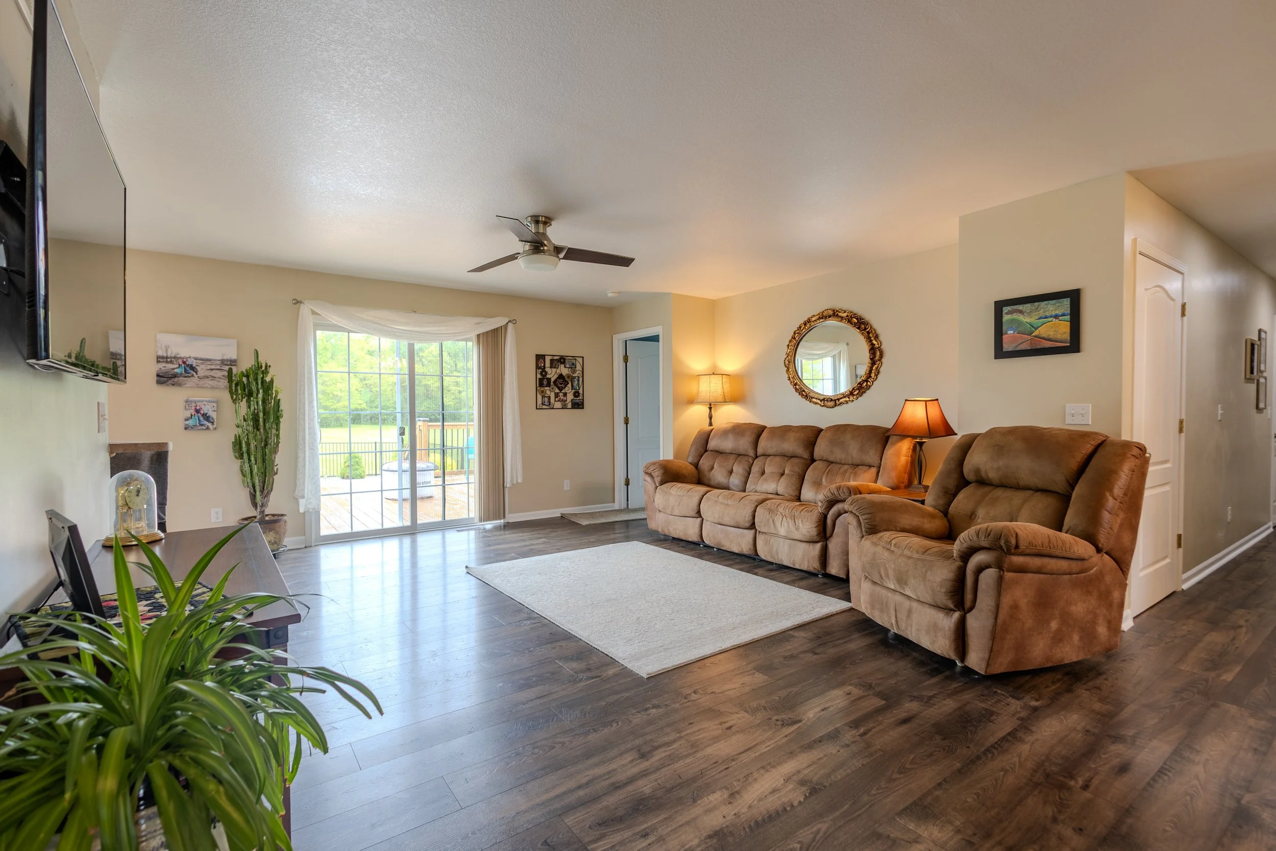 Living room with beige walls, a three-cushion beige sofa, matching recliner, wooden flooring, sliding glass door leading to outdoor area, wall-mounted TV, potted plants, framed pictures, and standing lamps.