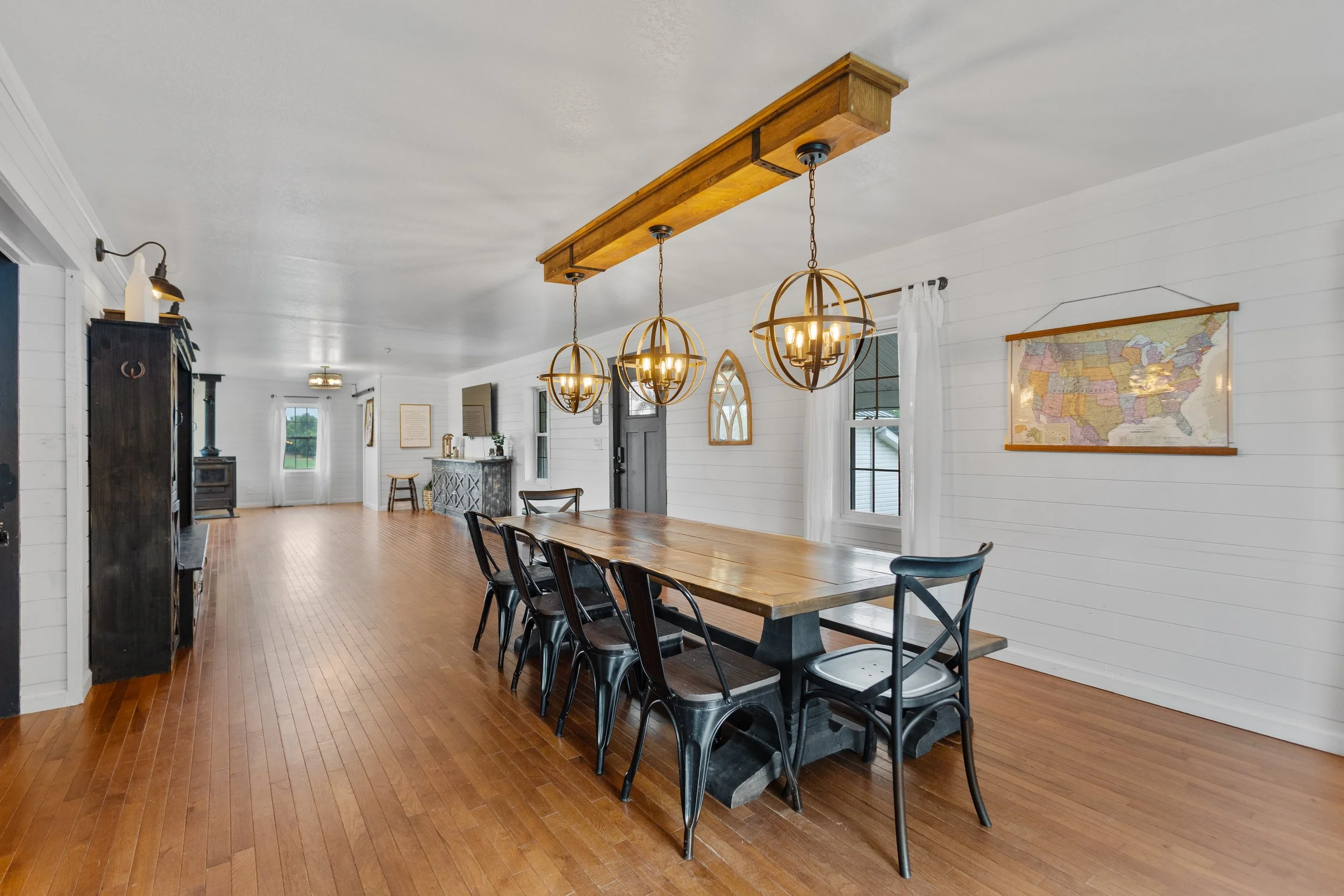 A spacious dining room with a large wooden table, six black chairs, and three spherical pendant lights hanging from a wooden beam. The room has white shiplap walls, hardwood flooring, and a wall map.
