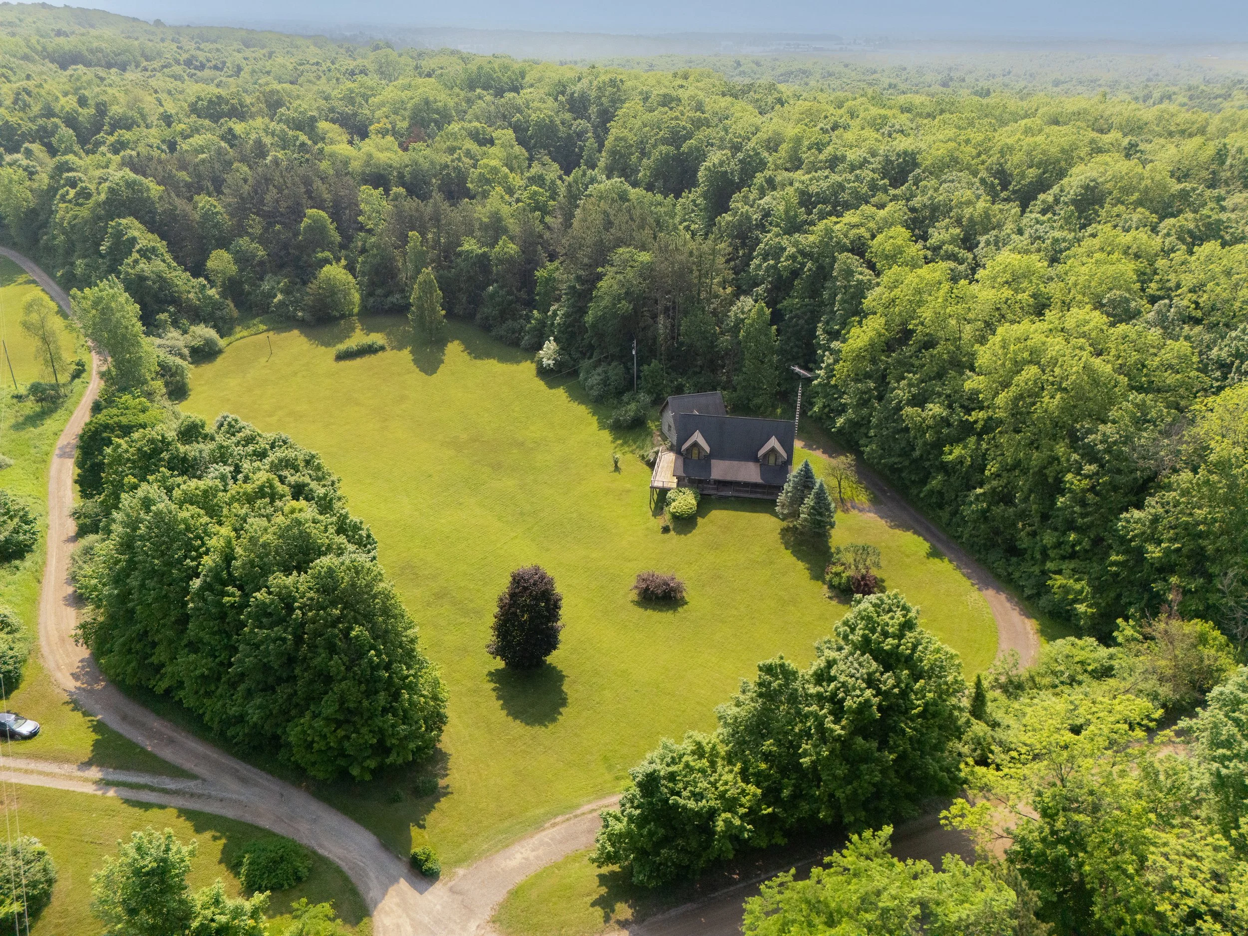 An aerial view of a large grassy lawn with a small house surrounded by trees and pathways, with a dense forest in the background.