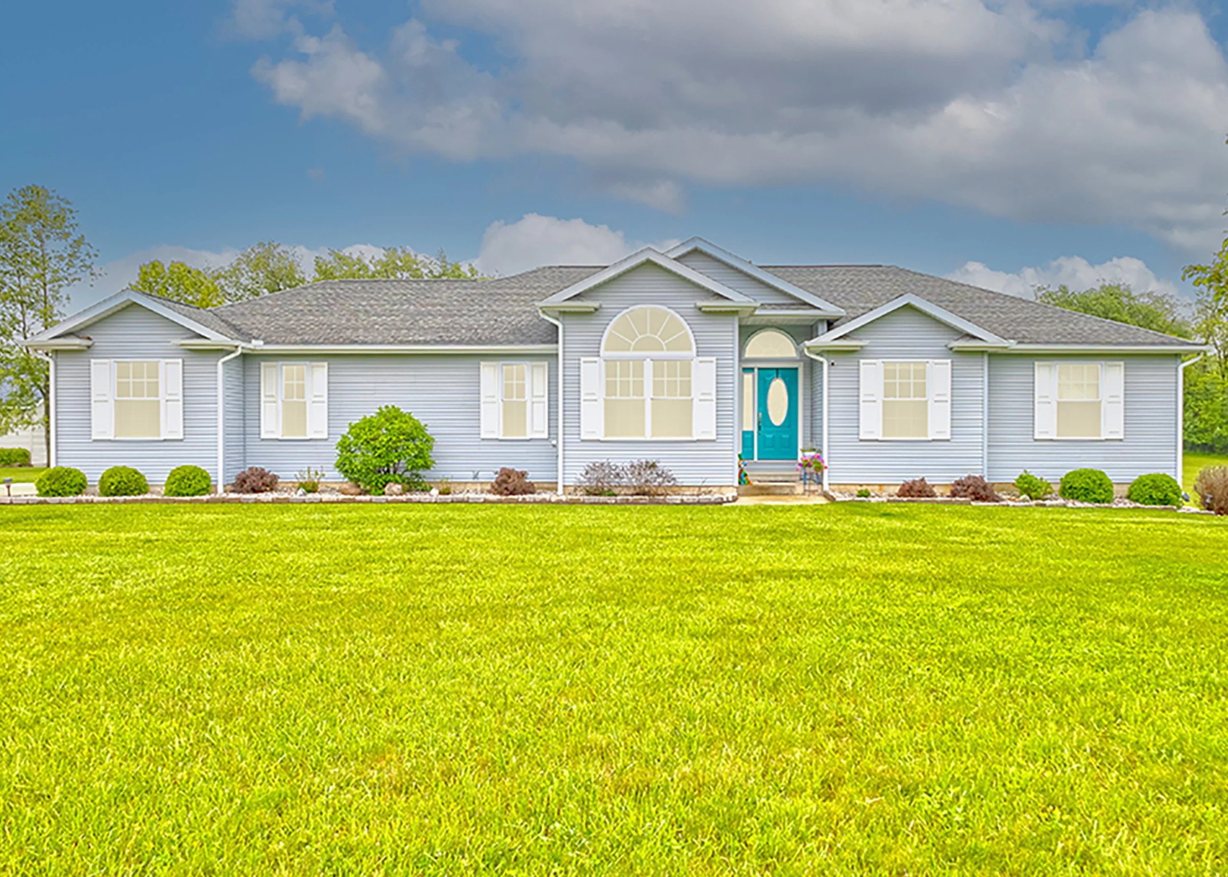 A large blue house with white shutters, surrounded by a green lawn and trees under a partly cloudy sky.