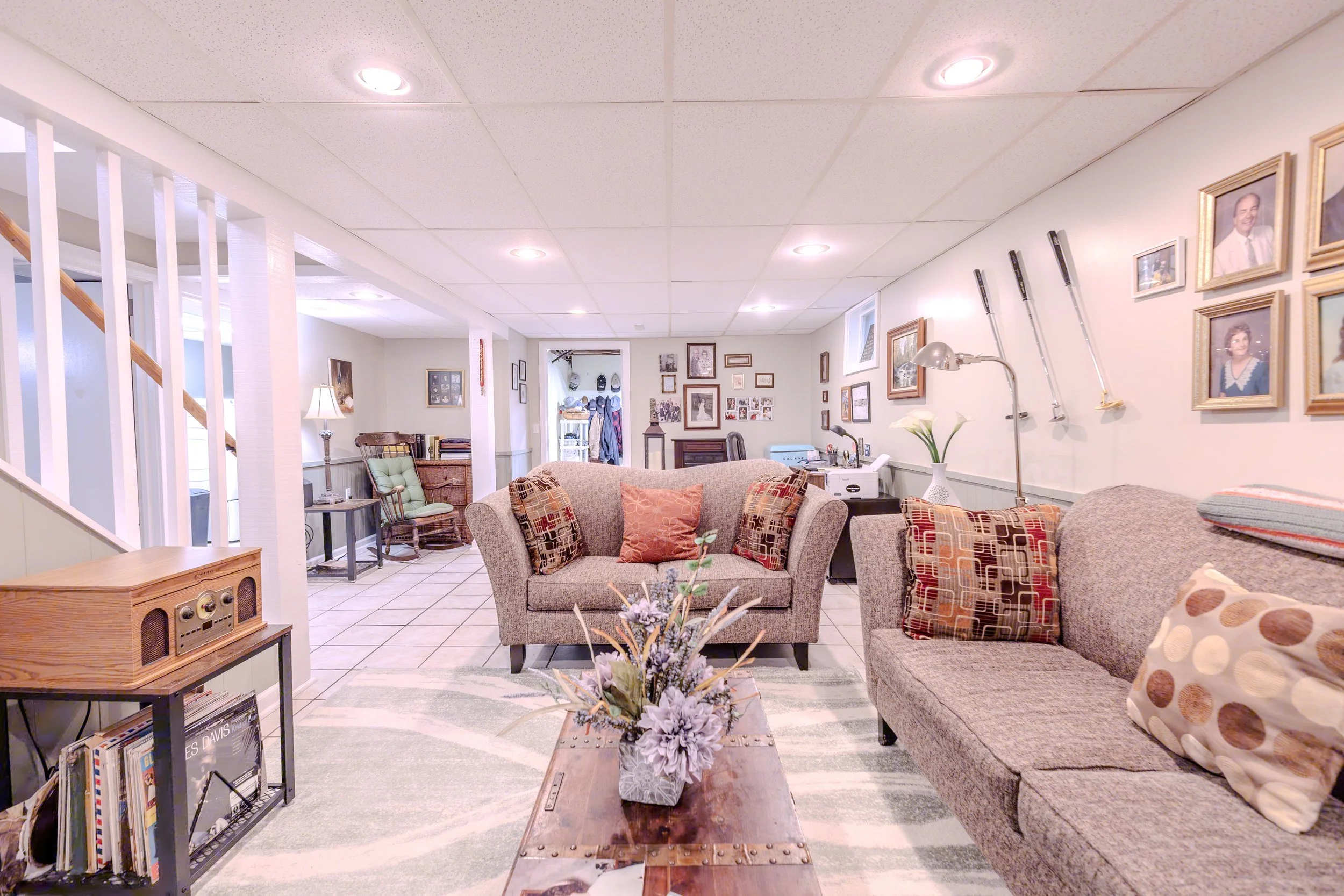 Living room with beige sofas, coffee table with a floral arrangement, gallery wall with framed photos, and a small sitting area with a lamp and bookshelf.