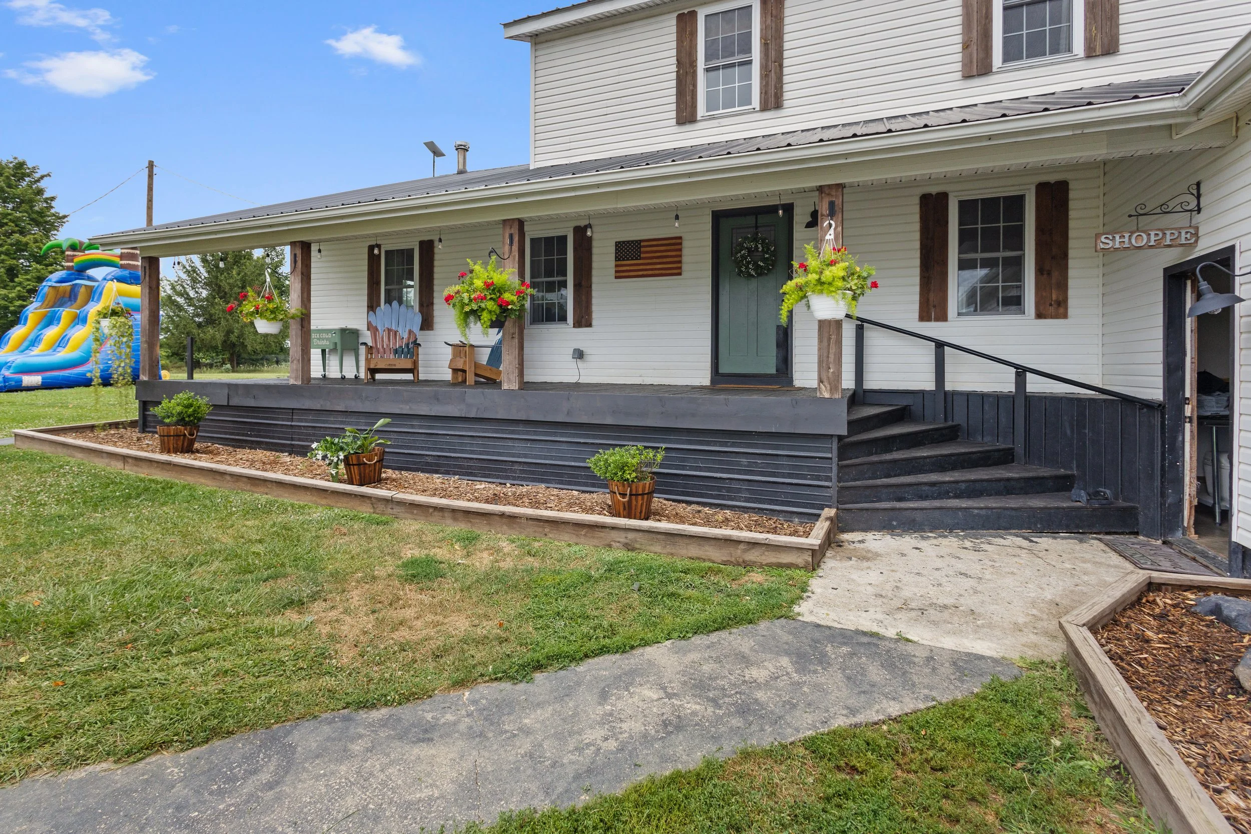 Front porch of a white house with wooden shutters, hanging flower baskets, Adirondack chairs, a wreath on the door, and outdoor decorations. There is a slide and a green patch in the yard.