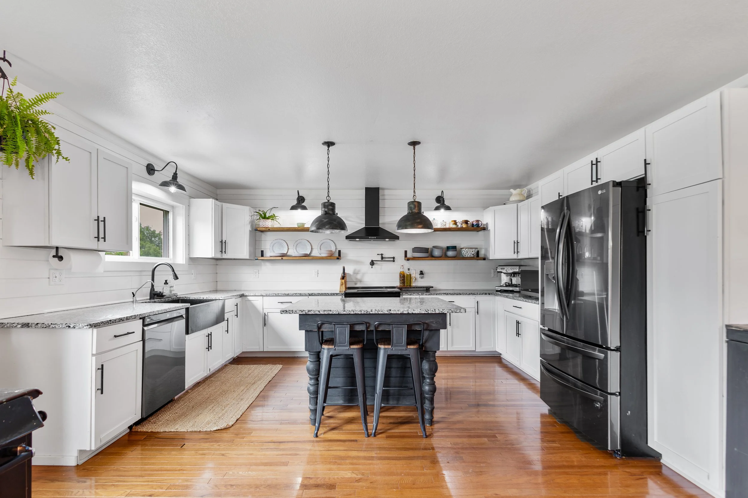 Modern kitchen with white cabinets, black hardware, black appliances including a refrigerator, and a black kitchen island with barstools. Open shelving with dishes and plants, hardwood floors, and three black pendant lights hanging above the island.