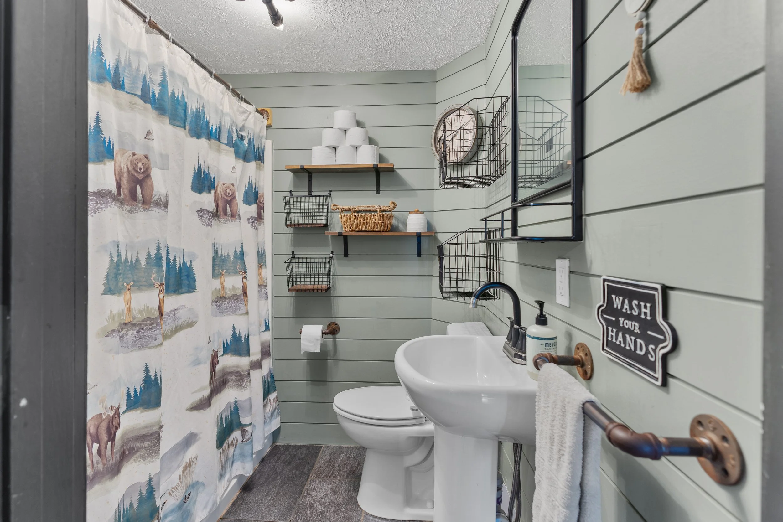 Bathroom with light green shiplap walls, a white toilet, a pedestal sink, a rustic towel bar, and a shower curtain featuring bears, deer, and trees.
