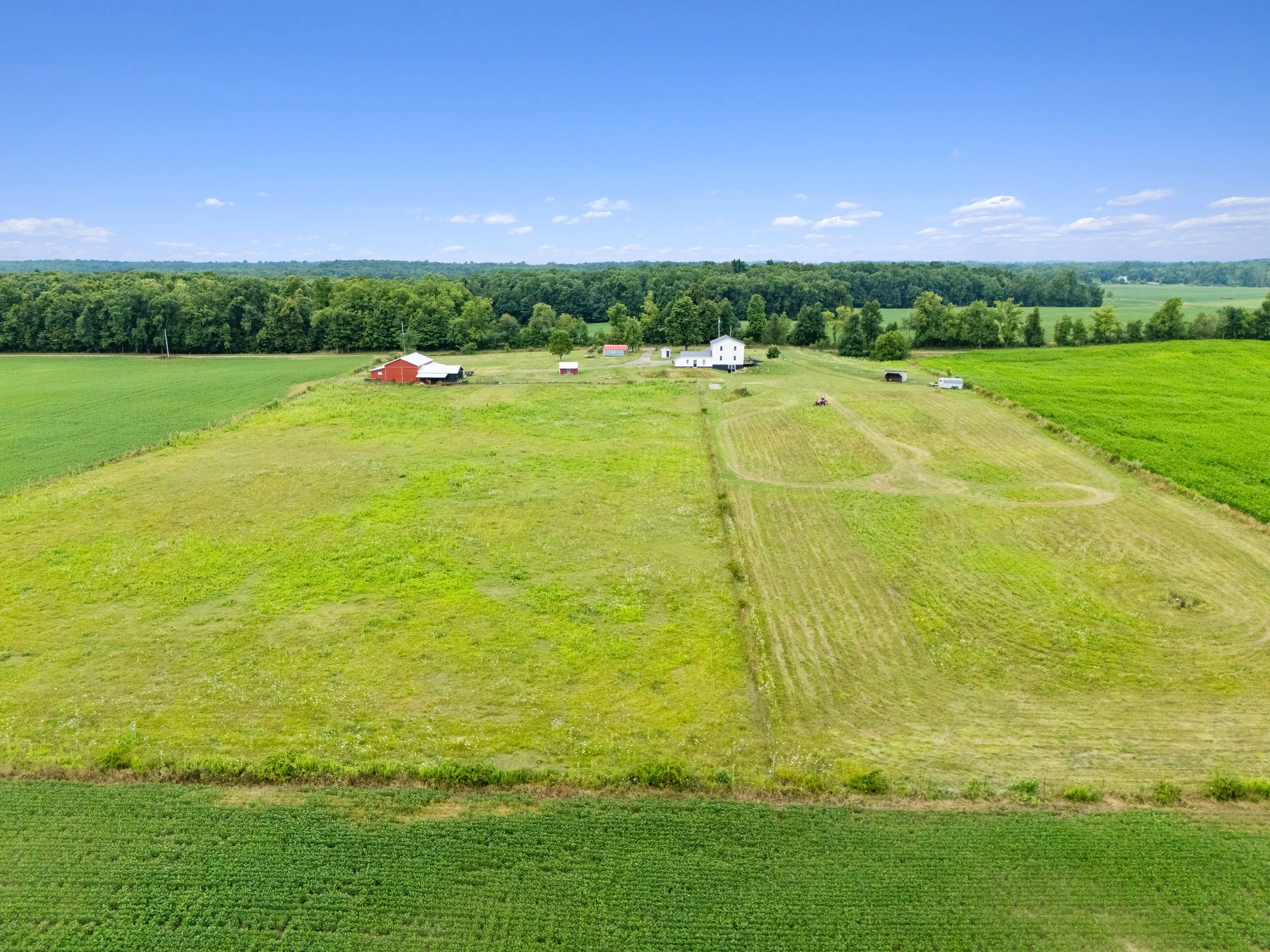 Aerial view of a large farm with a white barn, smaller red and white structures, and fields of grass and crops under a blue sky with scattered clouds.
