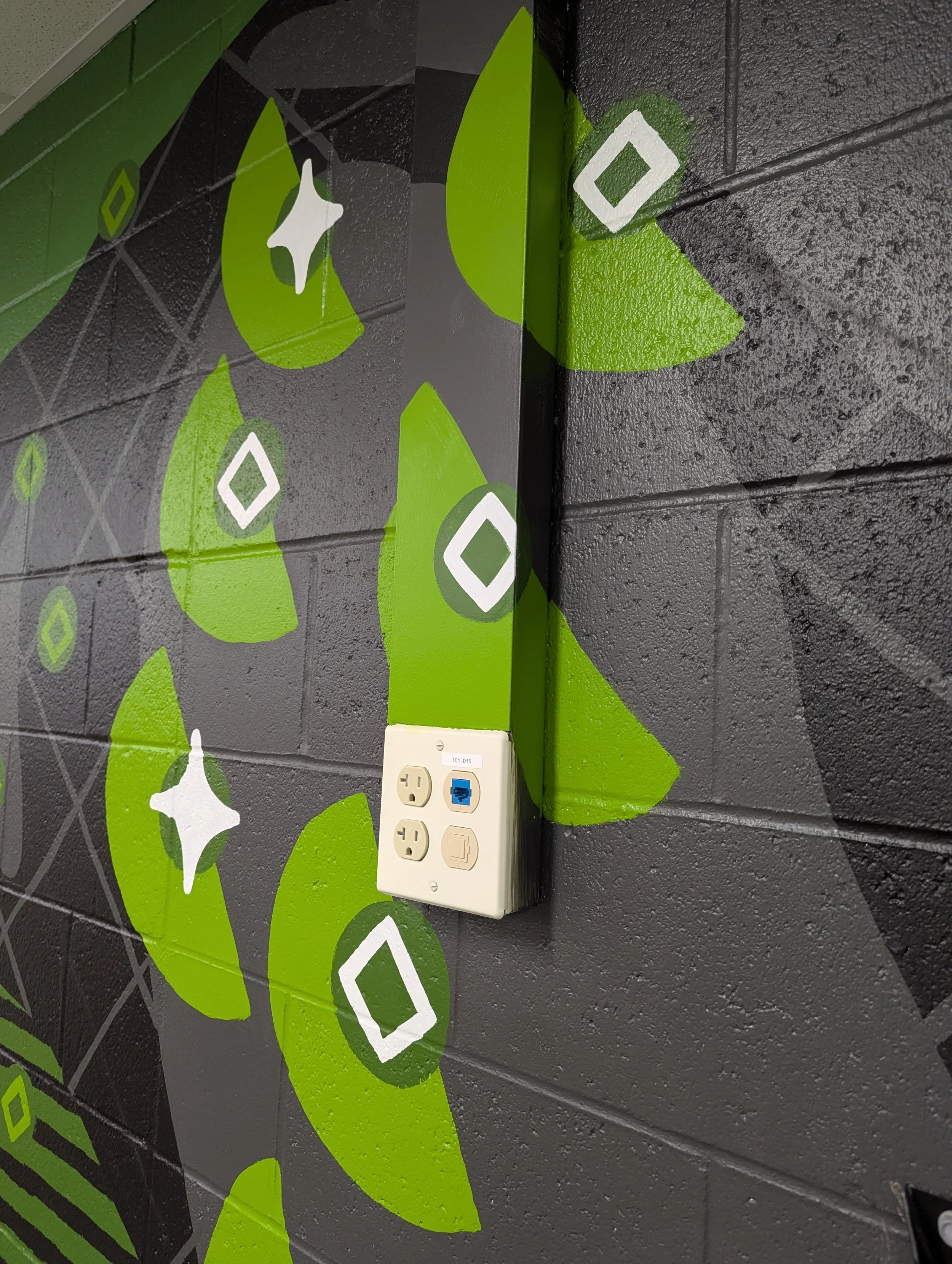 Angled close-up of painted geometric shapes surrounding an electrical box on the elementary school mural wall.