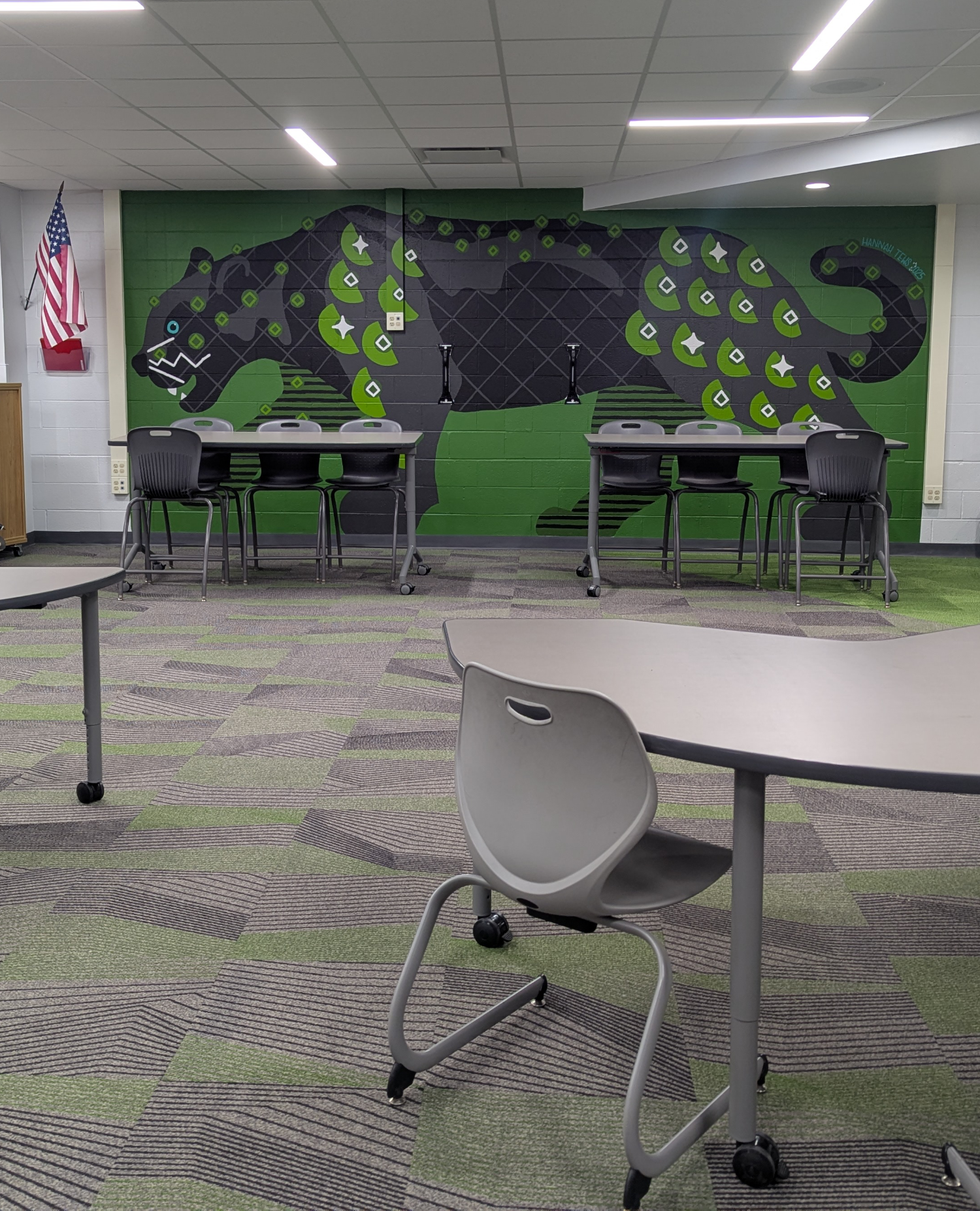 Empty classroom with tables and chairs, featuring a large mural of a black panther on the back wall and an American flag on the side wall.