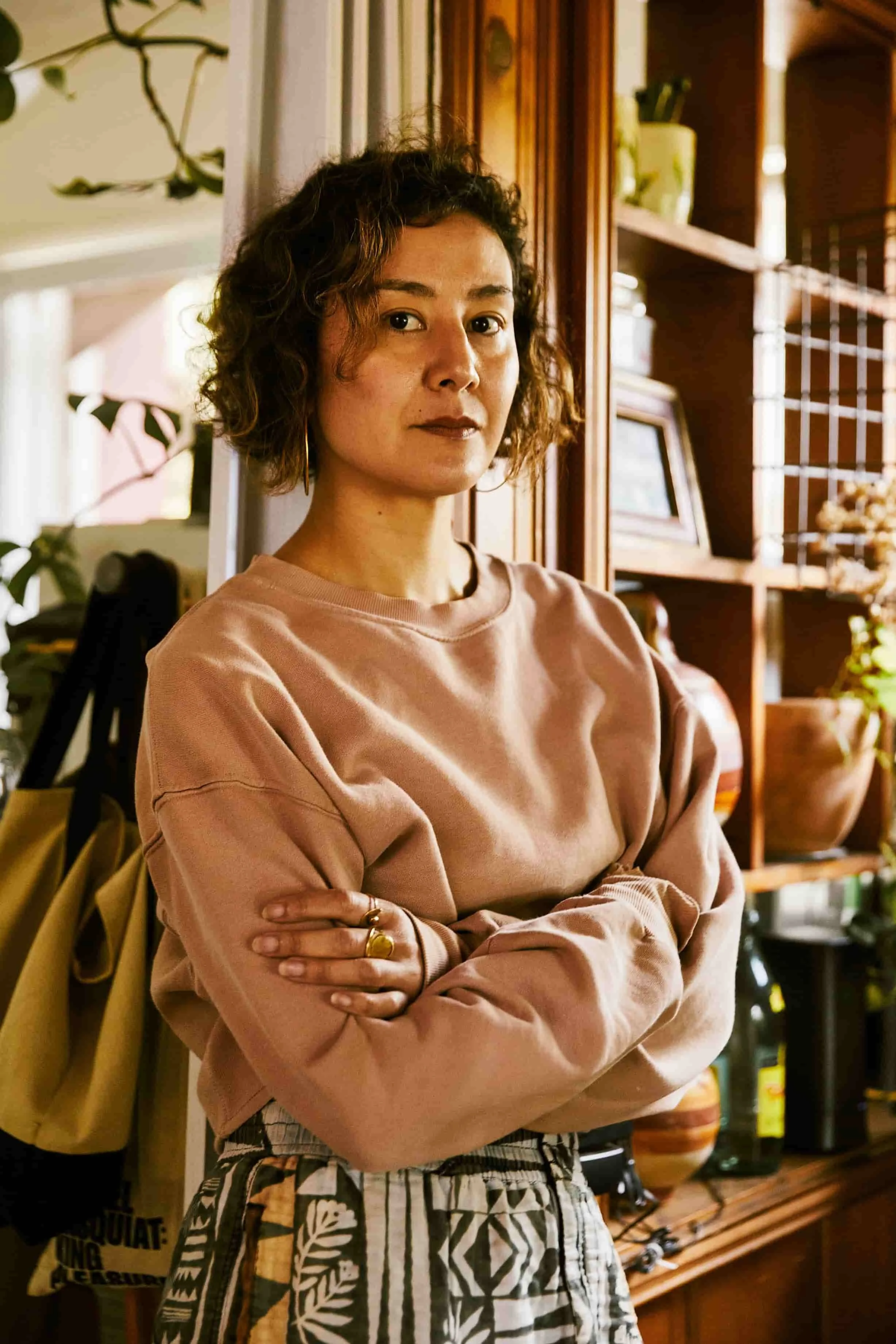 pilates teacher with brown curly hair wearing pink sweatsheet with arms folded and wooden shelves in the background