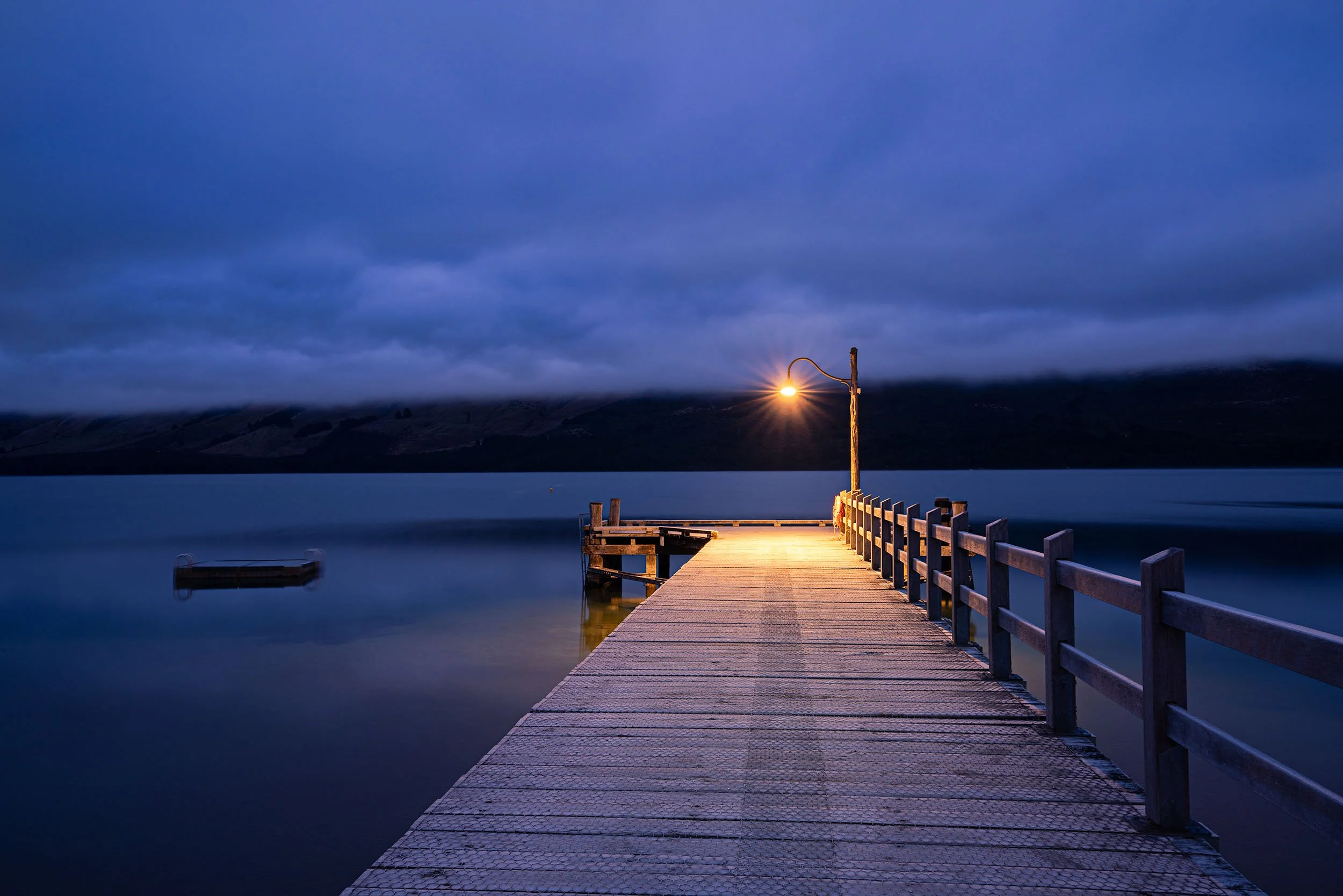 A wooden dock extending into a calm body of water at dusk, illuminated by a streetlamp at the end of the dock. The sky is cloudy and dimly lit, with mountains and a cloudy sky in the background.