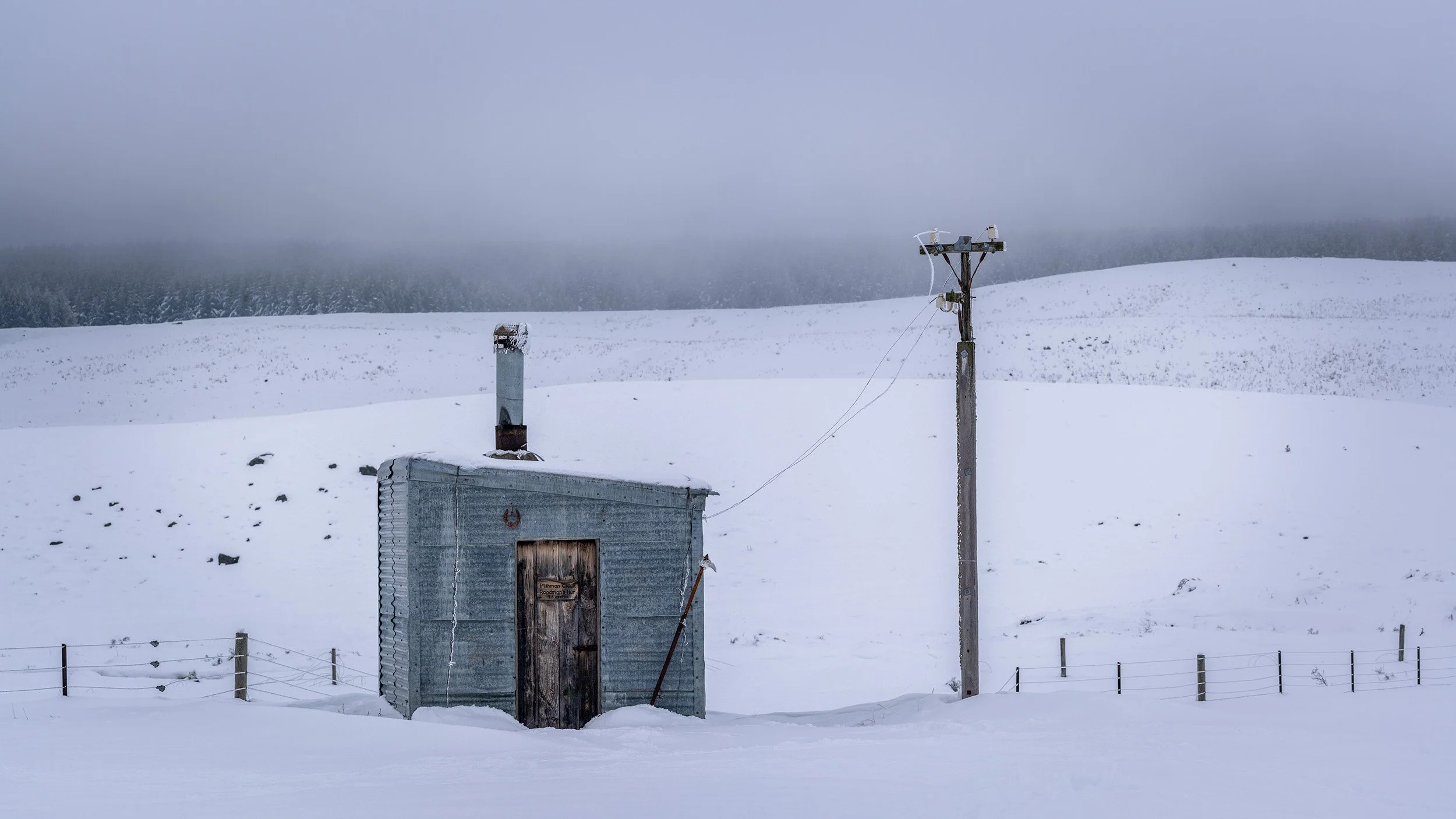 Snow-covered landscape with an old metal building, utility pole, and a snow fence in the foreground. Overcast sky and distant snowy hills and trees in the background.