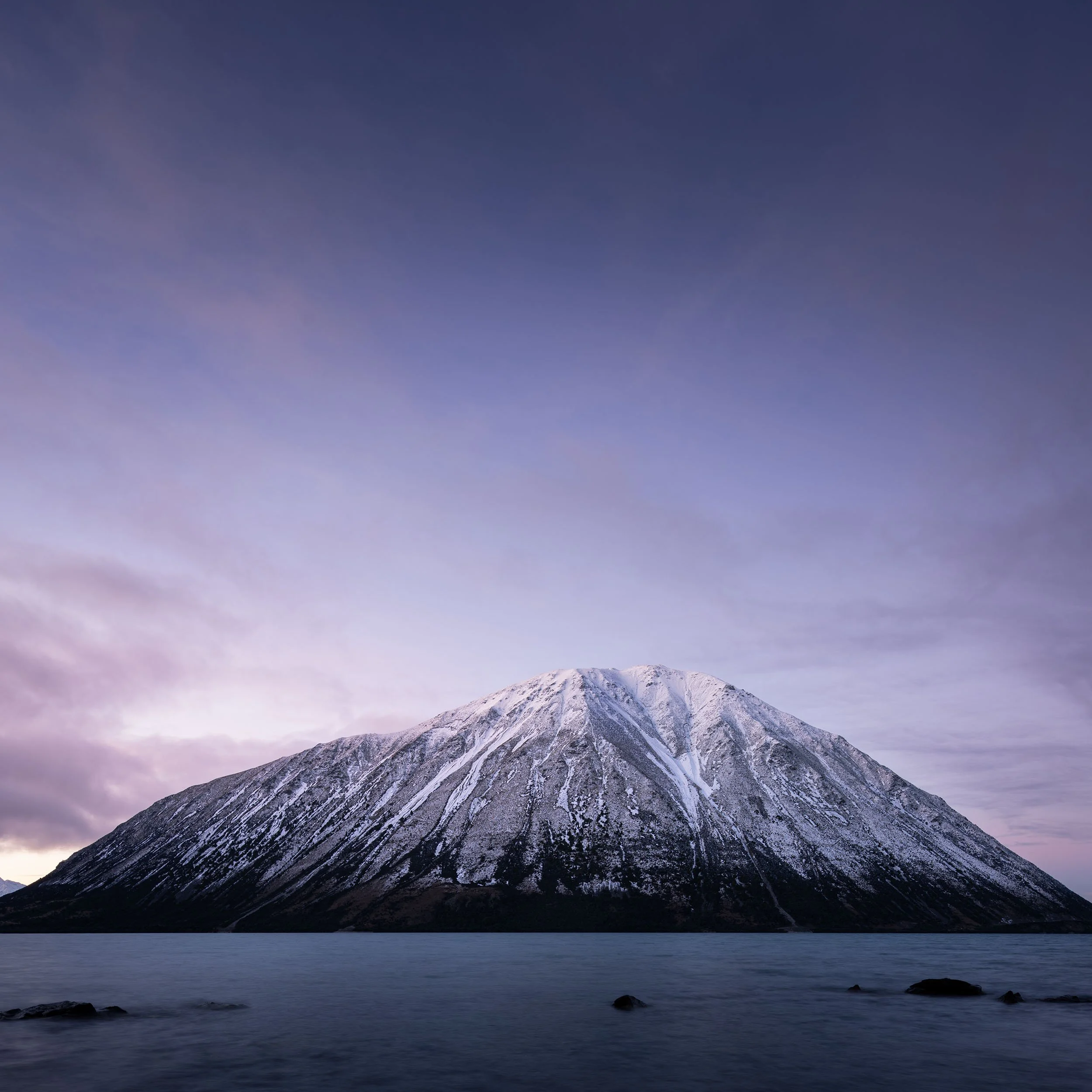 A snow-capped mountain rising above a body of water with rocks, under a partly cloudy sky at dusk or dawn.