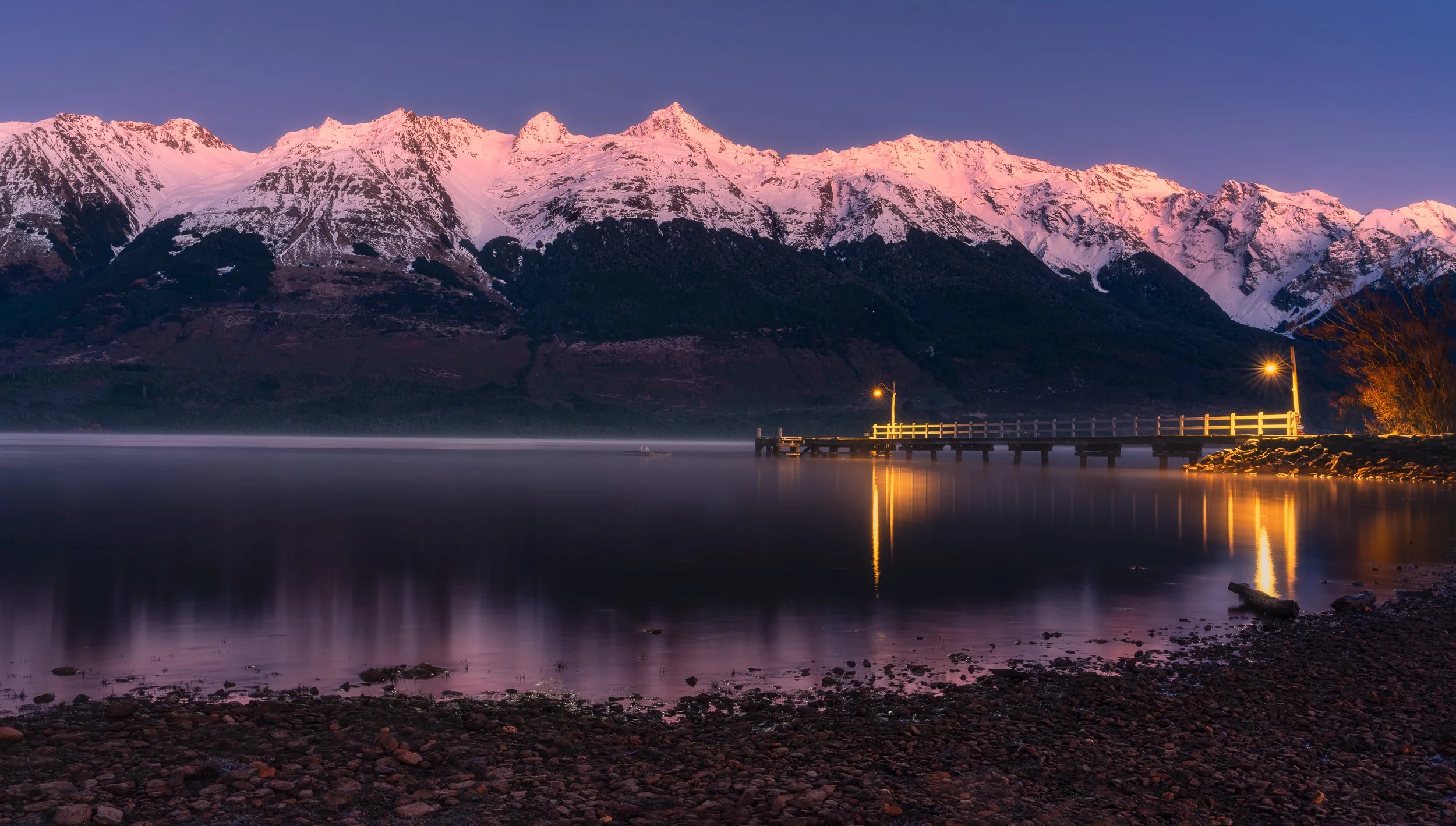 Snow-capped mountains during sunset over a calm lake with a dock and a small pier with street lamps, reflecting the pinkish light.