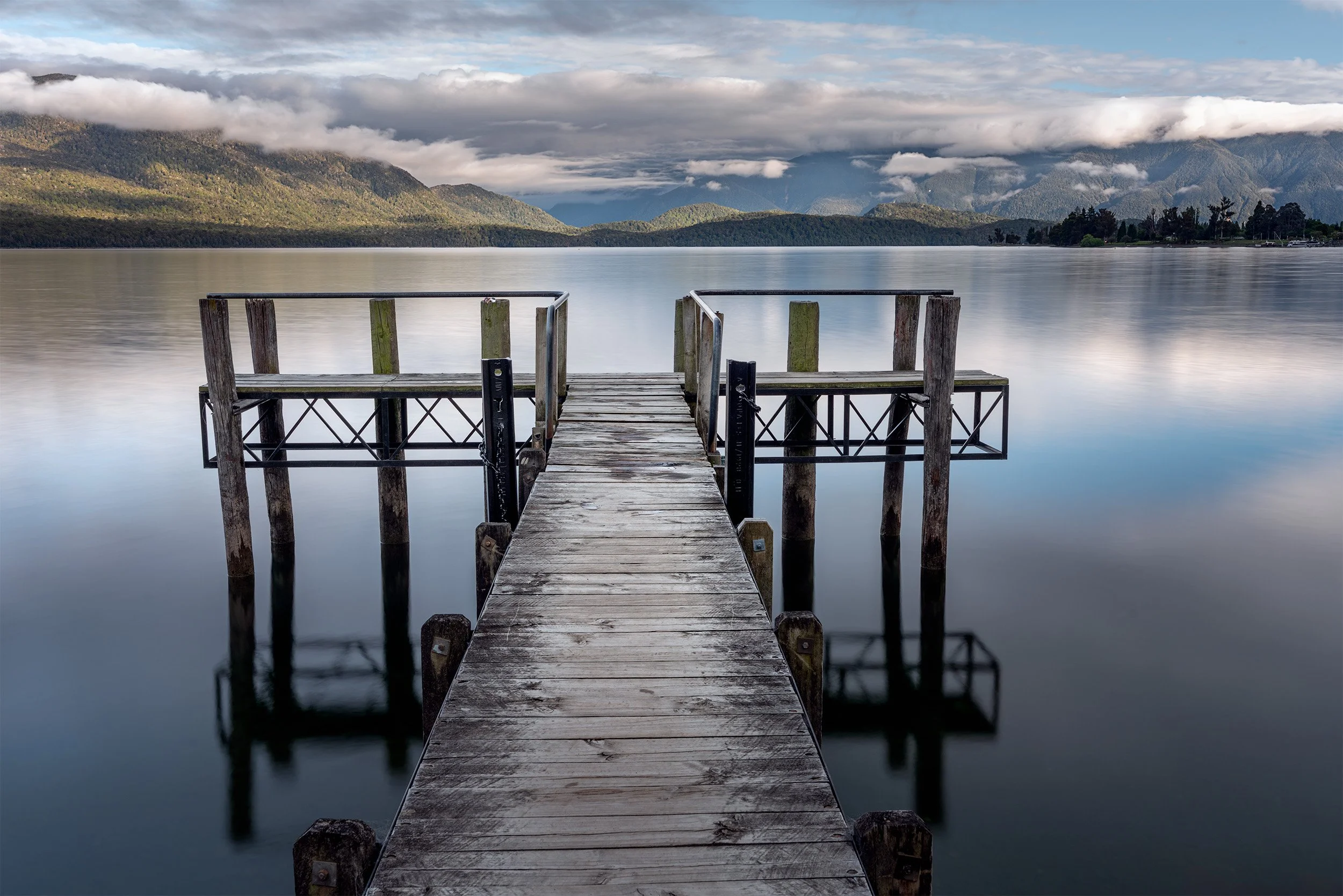 A wooden dock extends into a calm lake, with mountains and cloudy sky in the background.