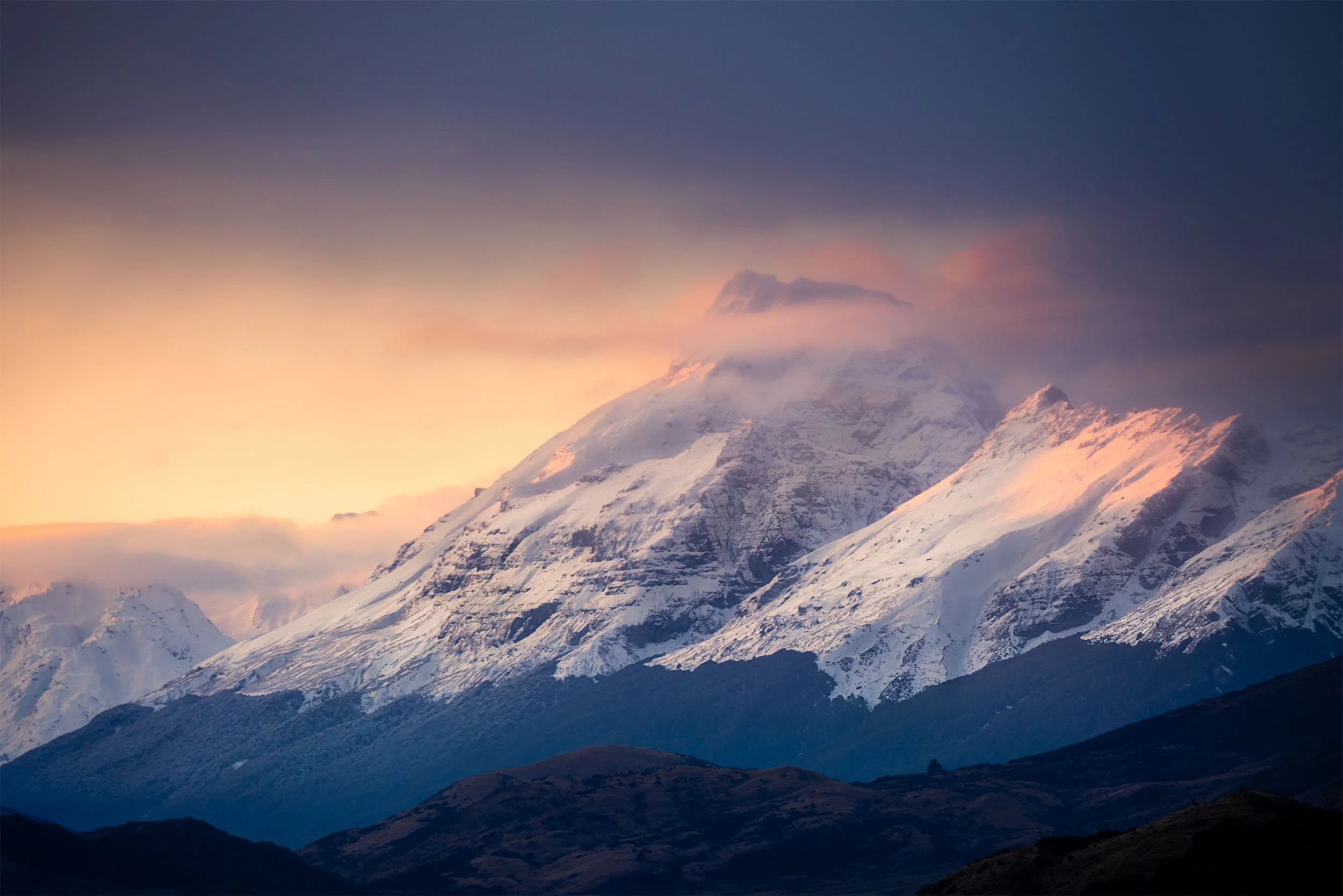 Snow-covered mountain range with a sunset sky and clouds at the top.