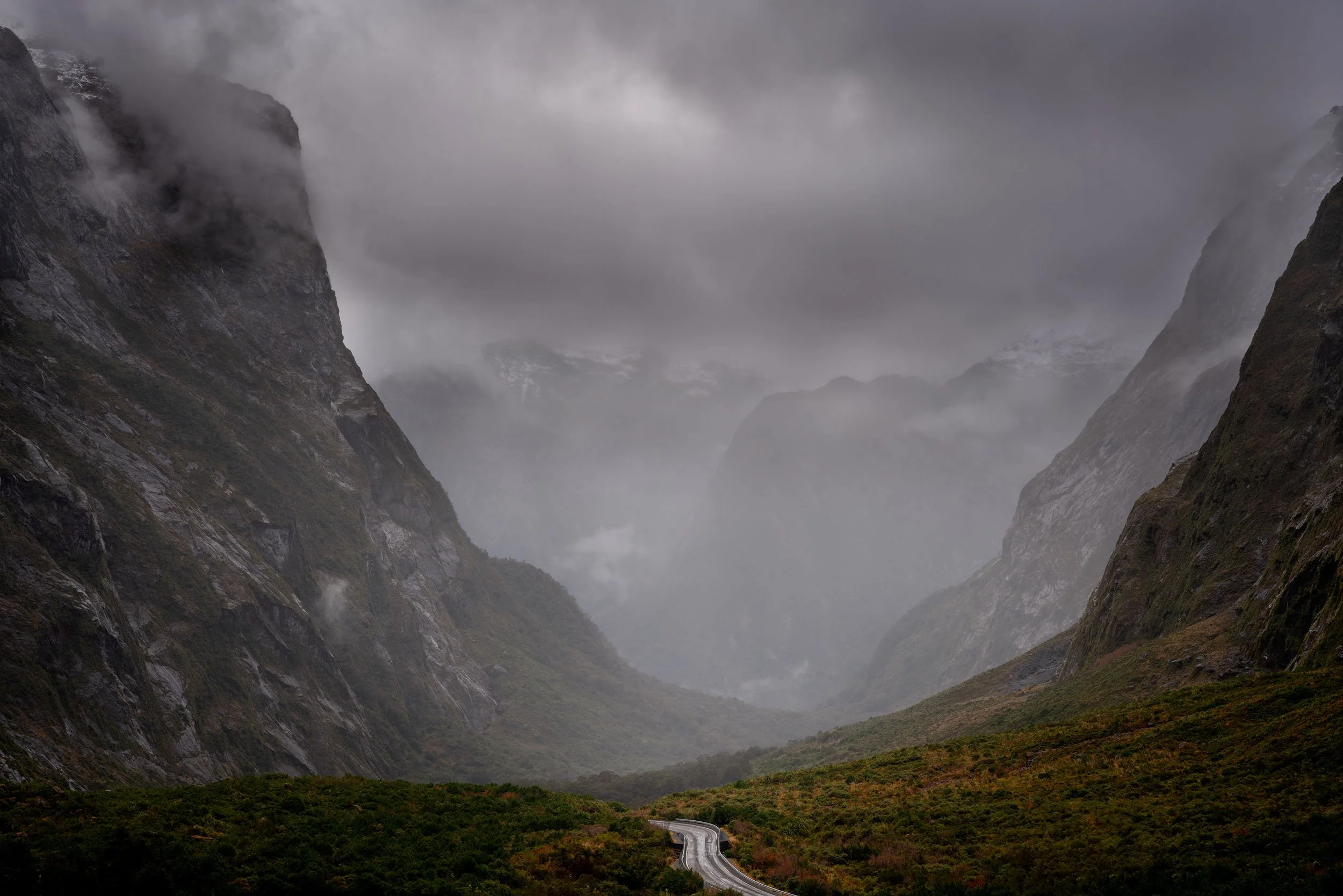 Foggy mountain valley with winding road, steep cliffs, and overcast sky.