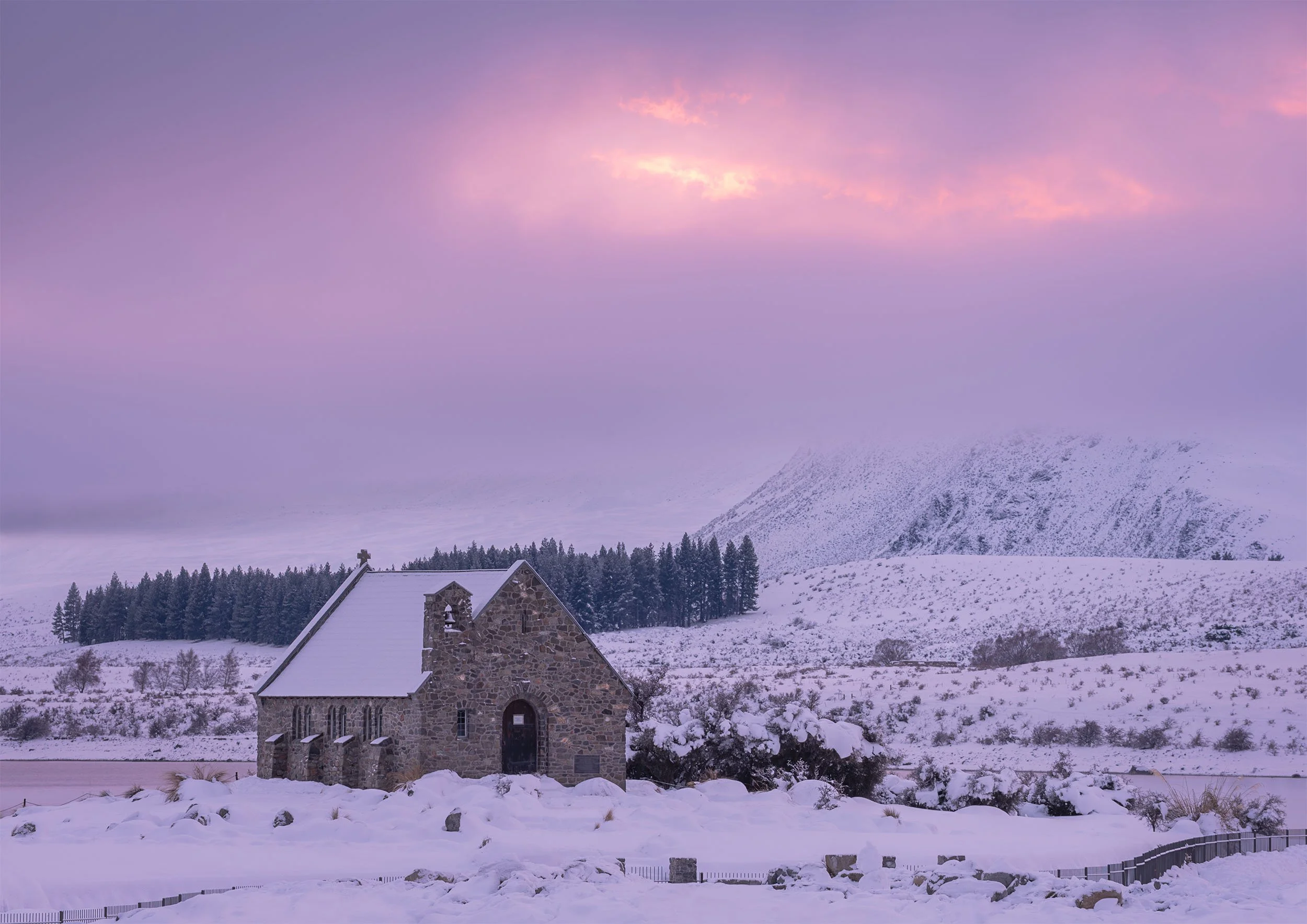 A snow-covered landscape with a small stone church in the foreground, surrounded by snow and trees, with a mountain in the background and a purple-pink sky.