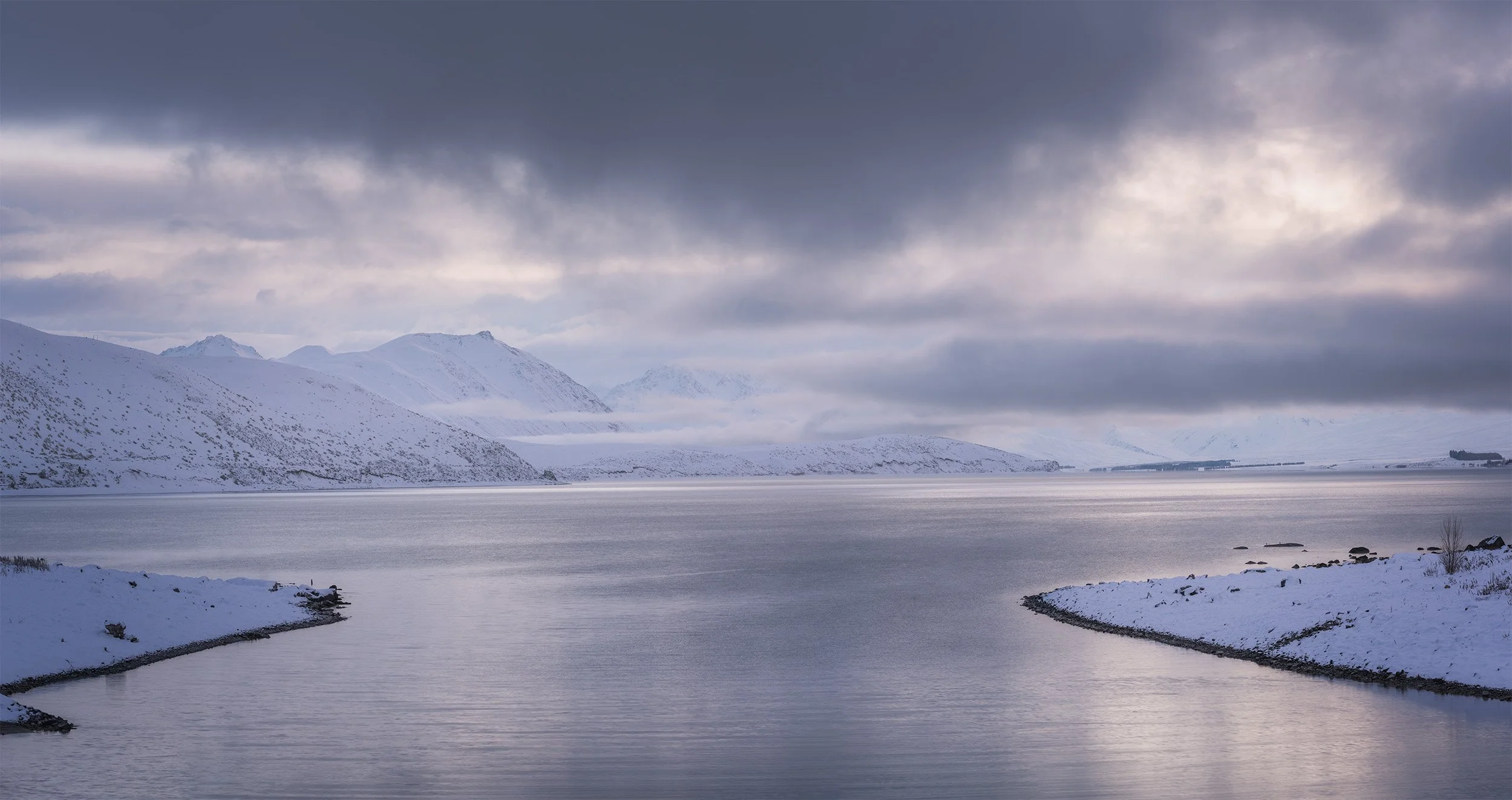 A snowy landscape with mountains in the background, a calm body of water in the foreground, and a cloudy sky overhead.