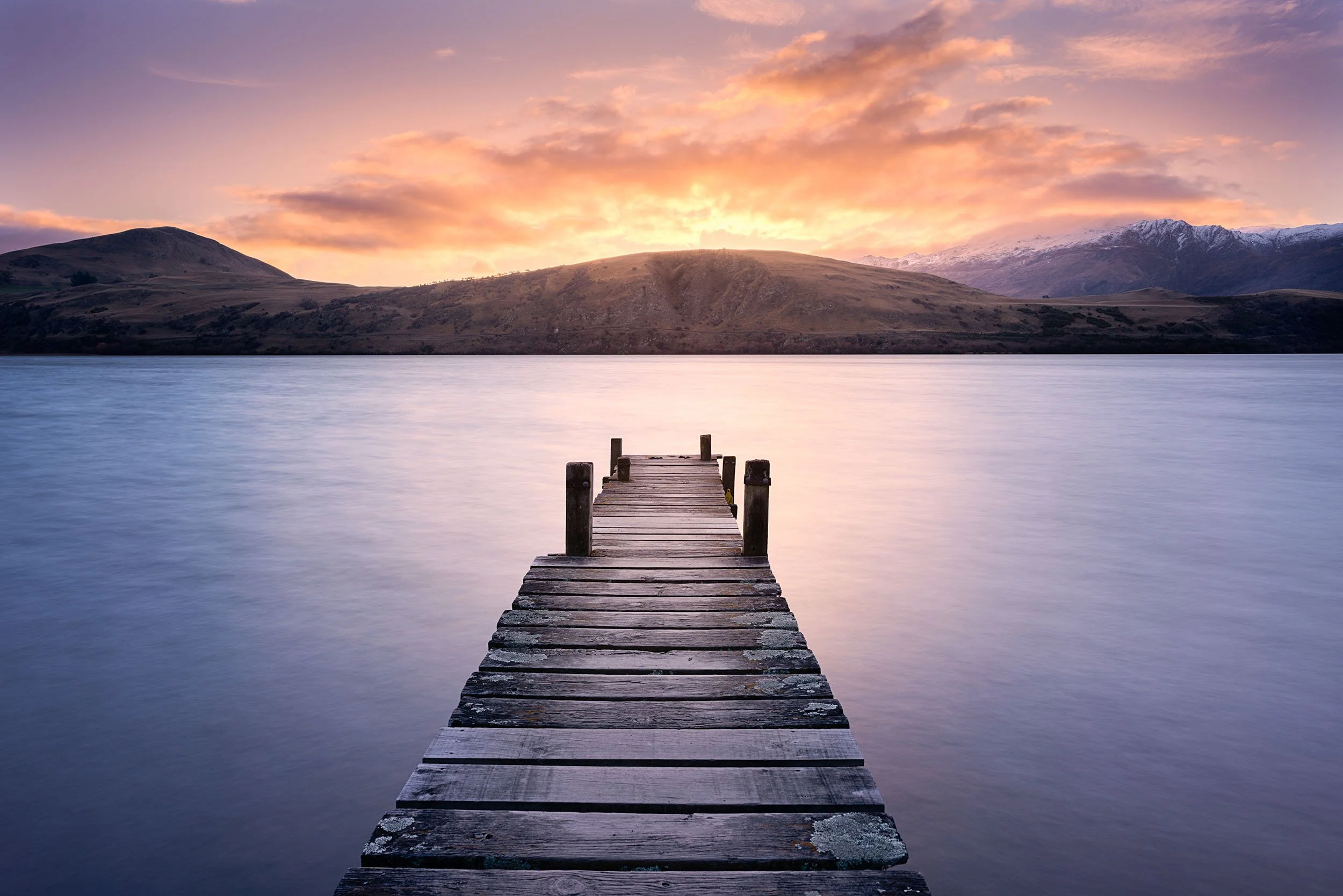 A wooden dock extends into a calm body of water with a hilly landscape and snow-capped mountains in the background, under a colorful sky during sunset or sunrise.