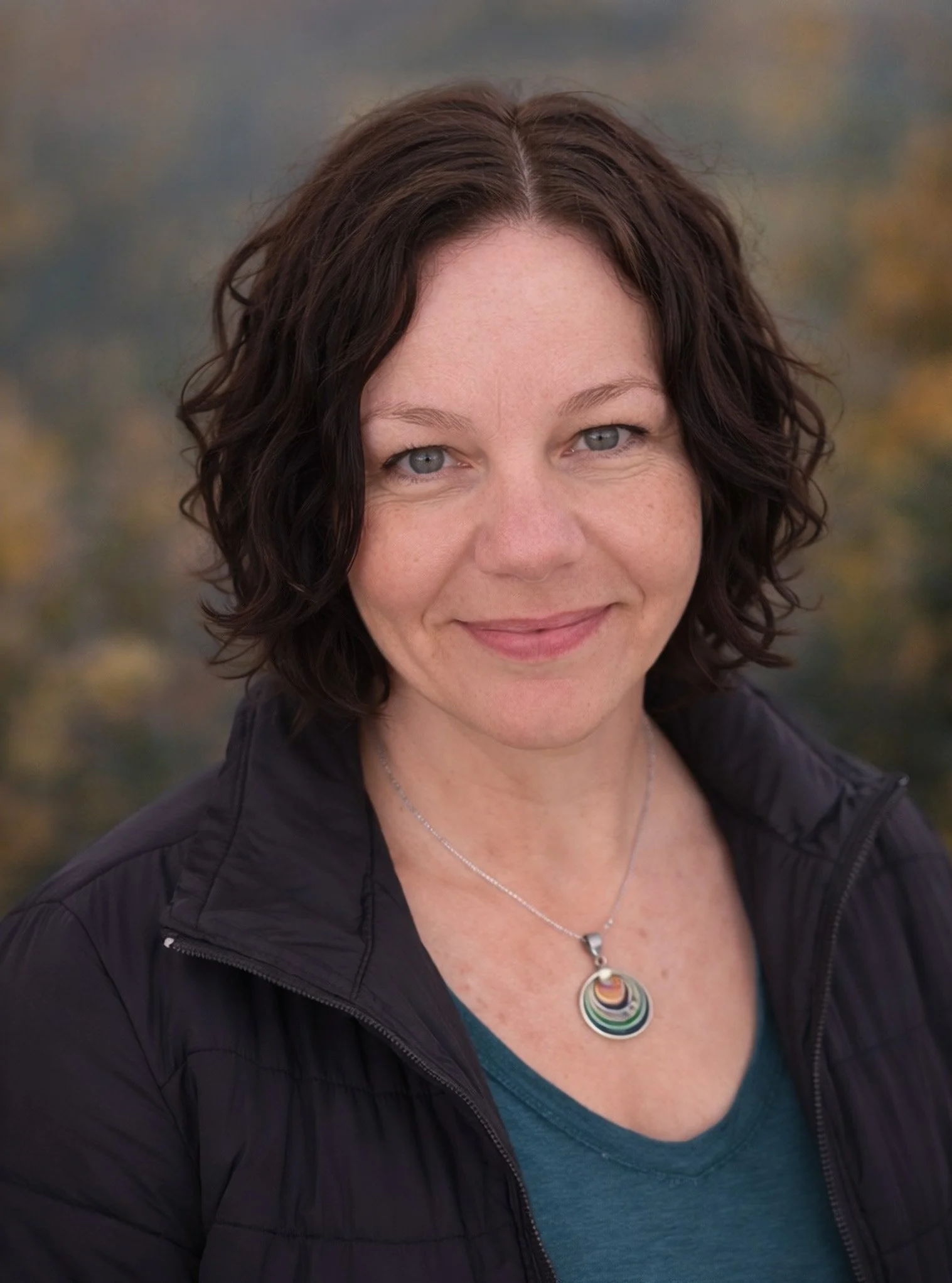 A woman with curly dark hair smiling outdoors with a blurred background of fall foliage, wearing a black jacket and a colorful circular pendant necklace.