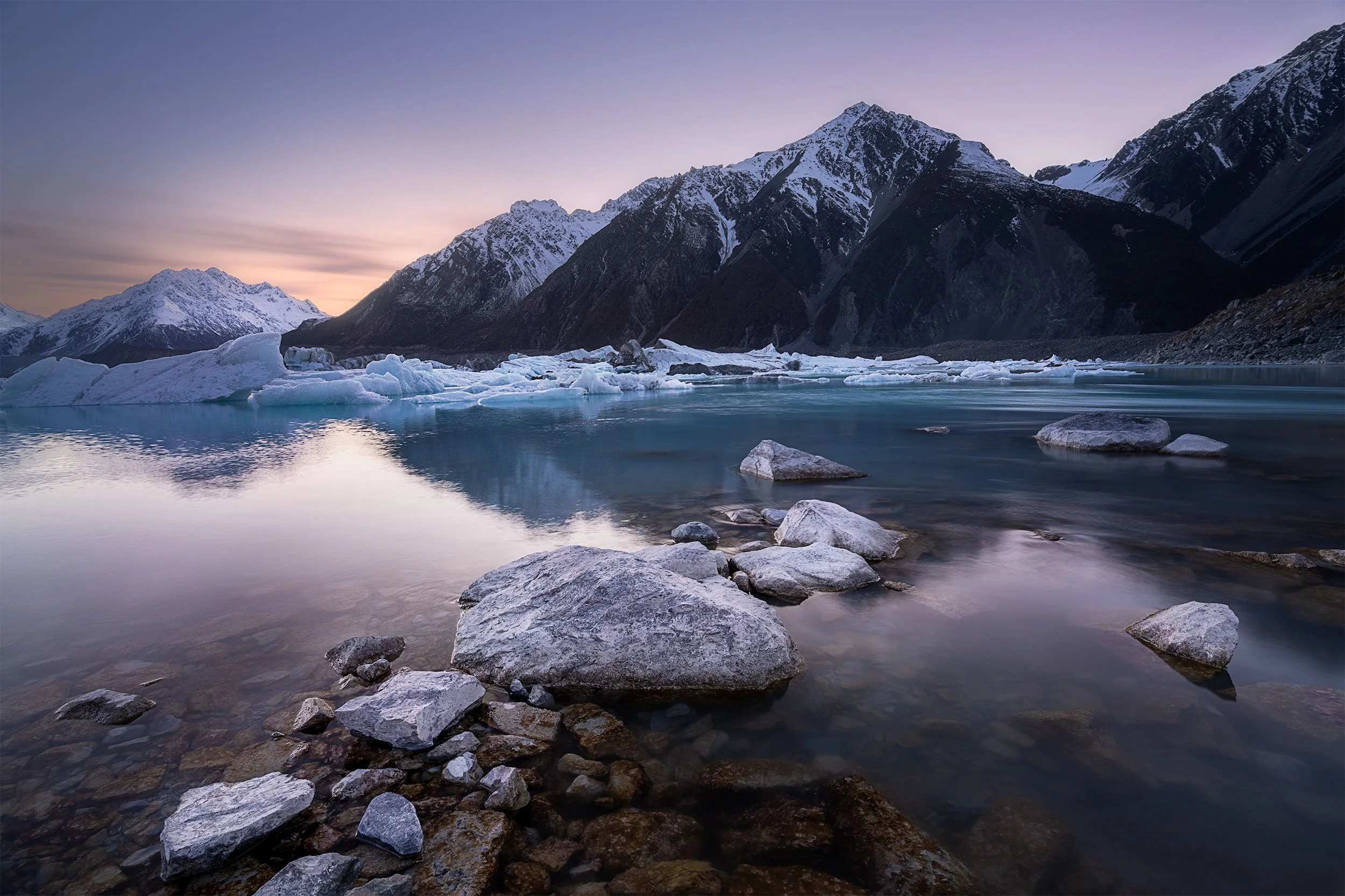 Snow-capped mountains reflected in a calm, icy blue lake with icebergs and rocks in the foreground during dusk.