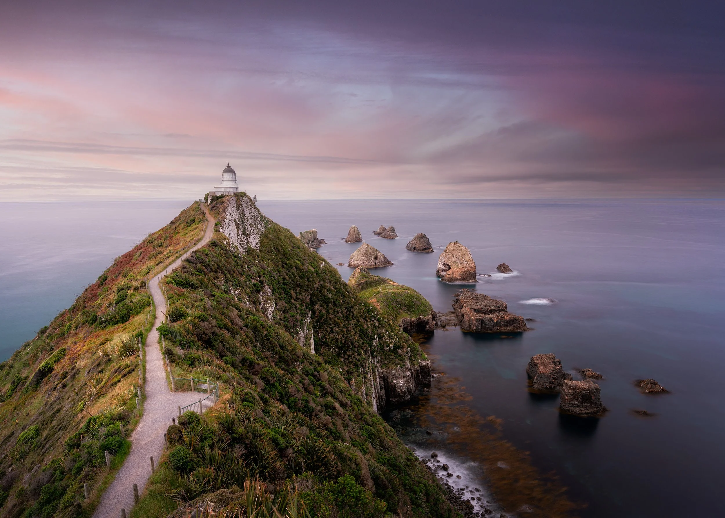 A lighthouse perched on a green, grassy hill at the end of a narrow, winding pathway along a rocky coast, with a calm sea and a cloudy, purple-tinted sky in the background.