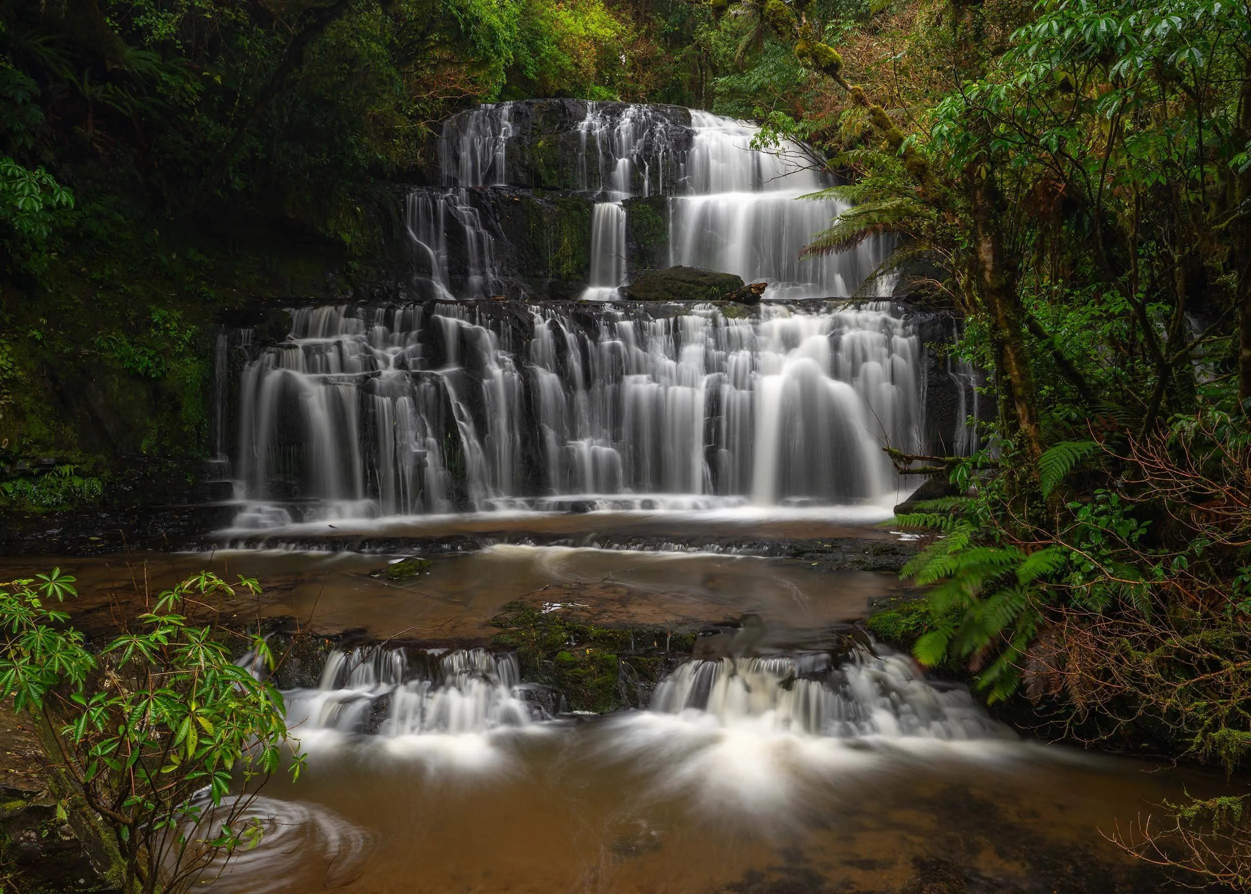 A multi-tiered waterfall flowing over rocks surrounded by lush green foliage in a dense forest.