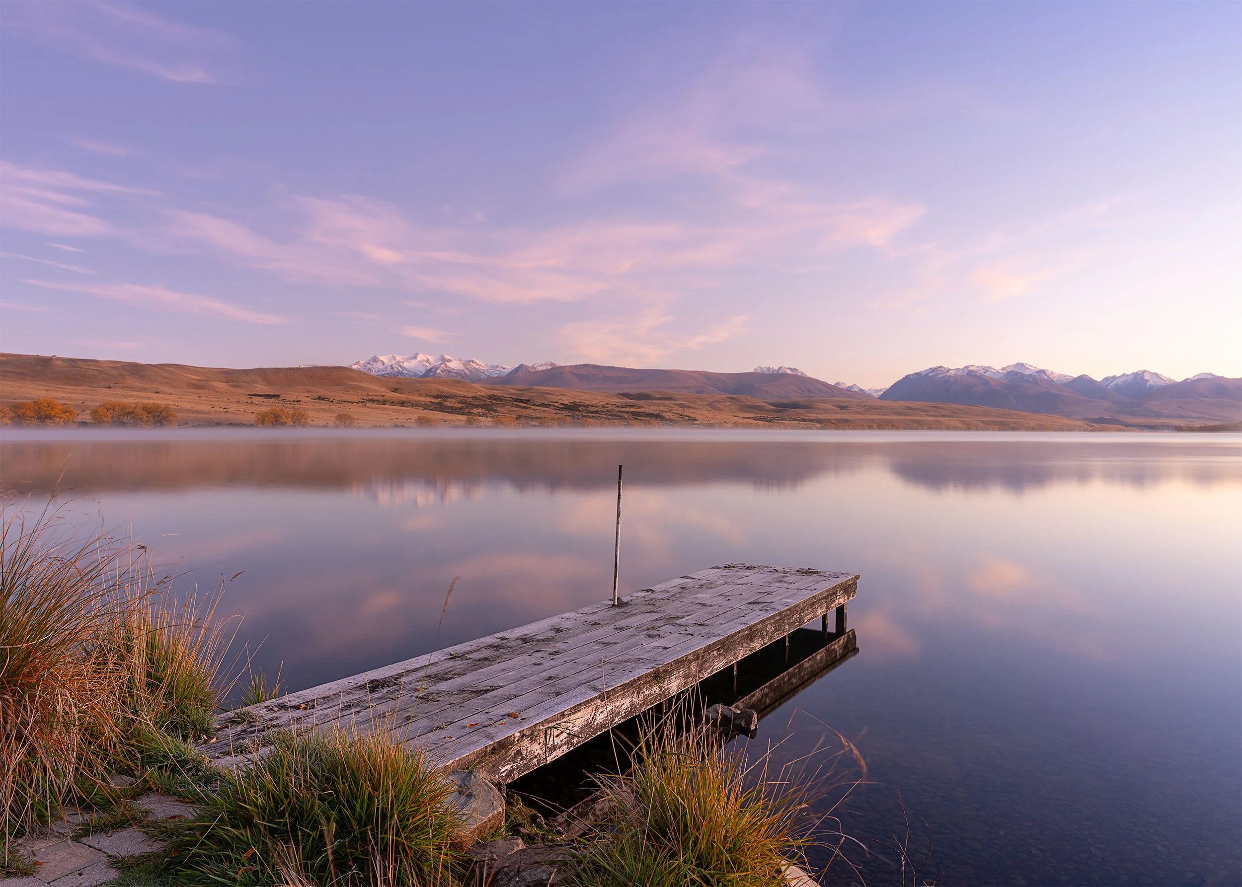 A calm lake with a wooden dock and a railing, surrounded by grassy banks, reflecting the sky and distant snow-capped mountains during sunset.