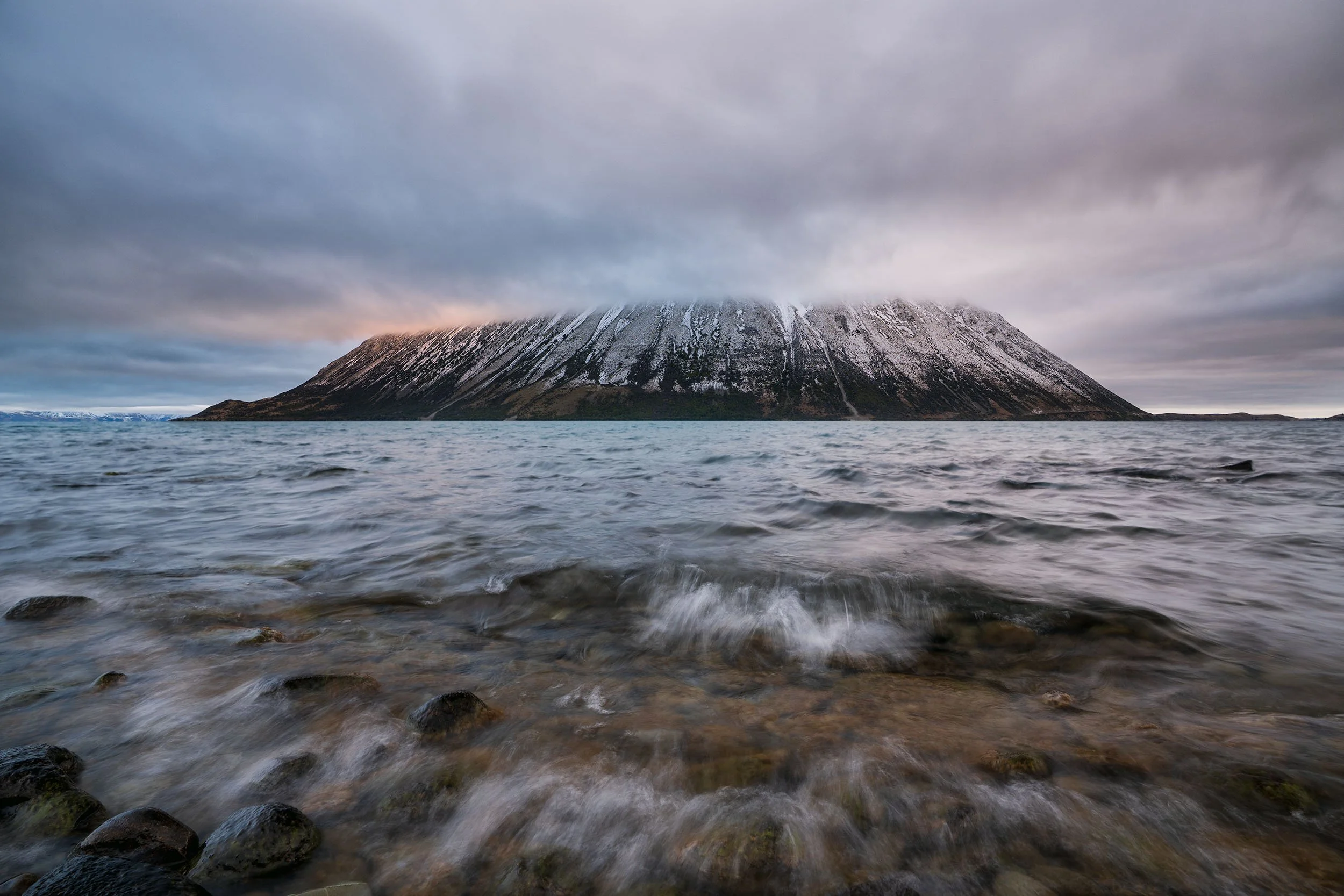 A large mountain with snow patches on its slopes, partially covered by clouds, surrounded by water with rocks in the foreground.