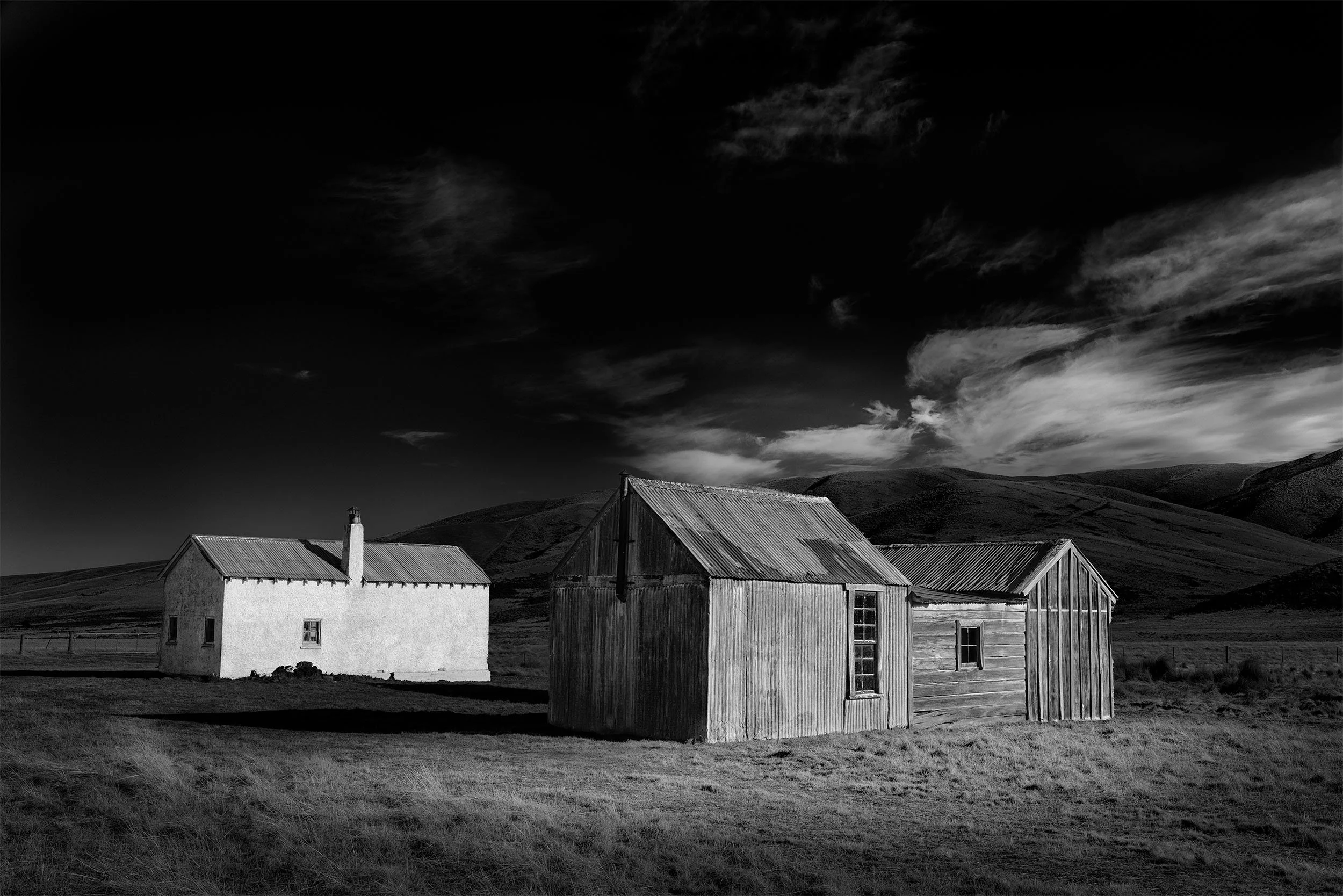 Black and white photo of four rustic farm buildings with hills and dramatic cloudy sky in the background.