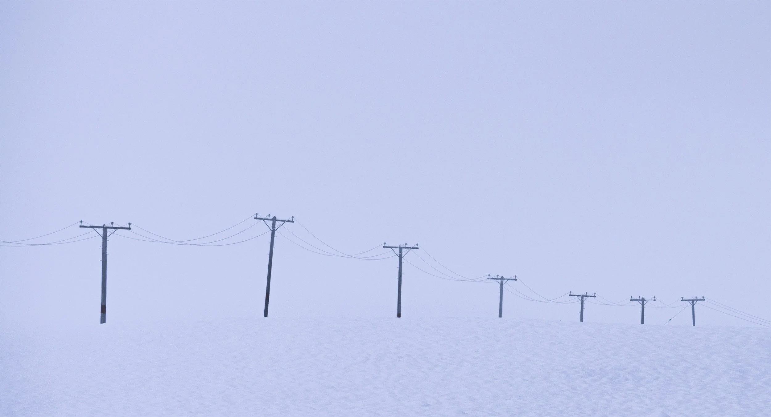 Line of utility poles with power lines against a gray, overcast sky, stretching across a snow-covered flat landscape.