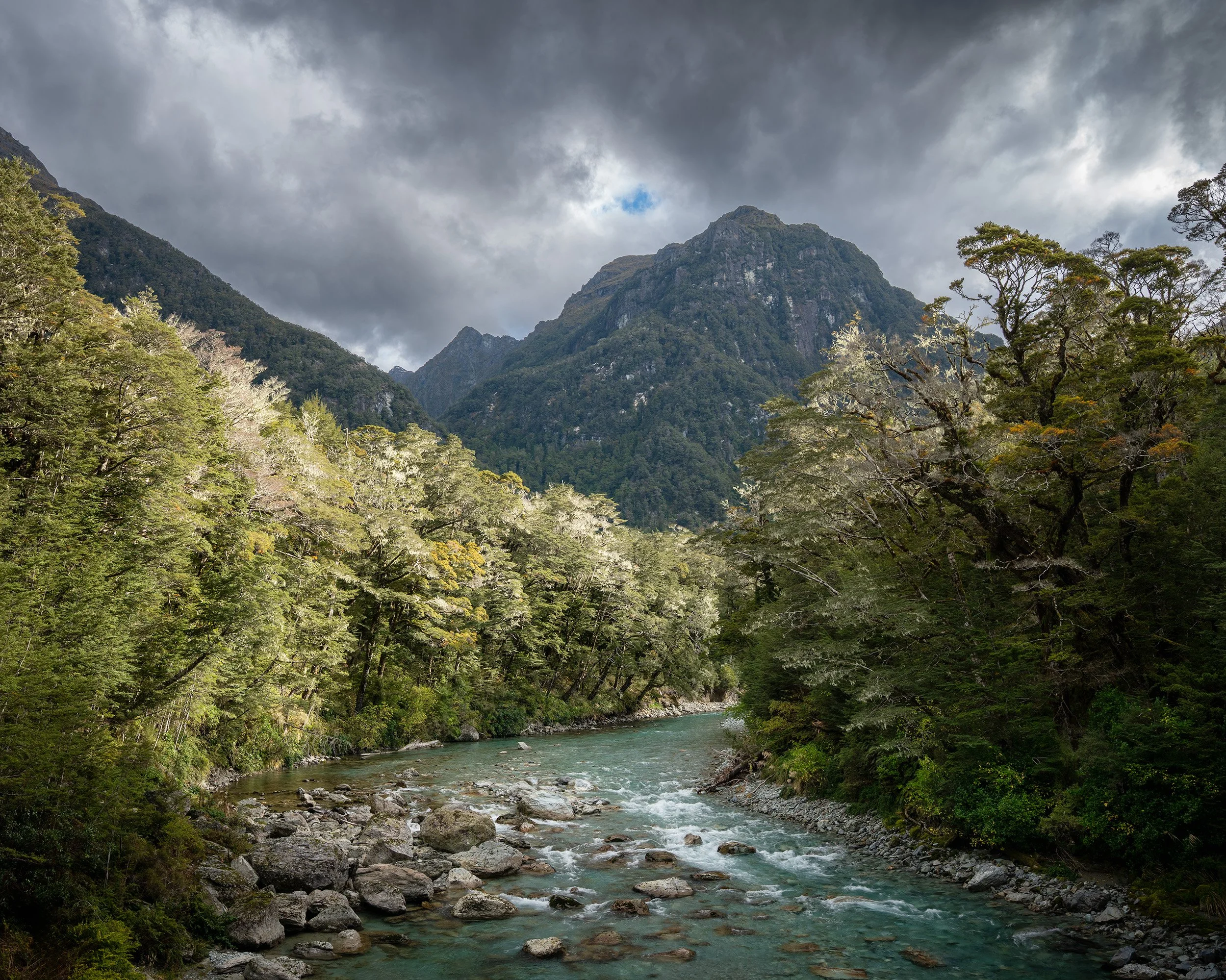 A mountain landscape featuring a river flowing through a forested valley surrounded by steep, rugged mountains under a cloudy sky.
