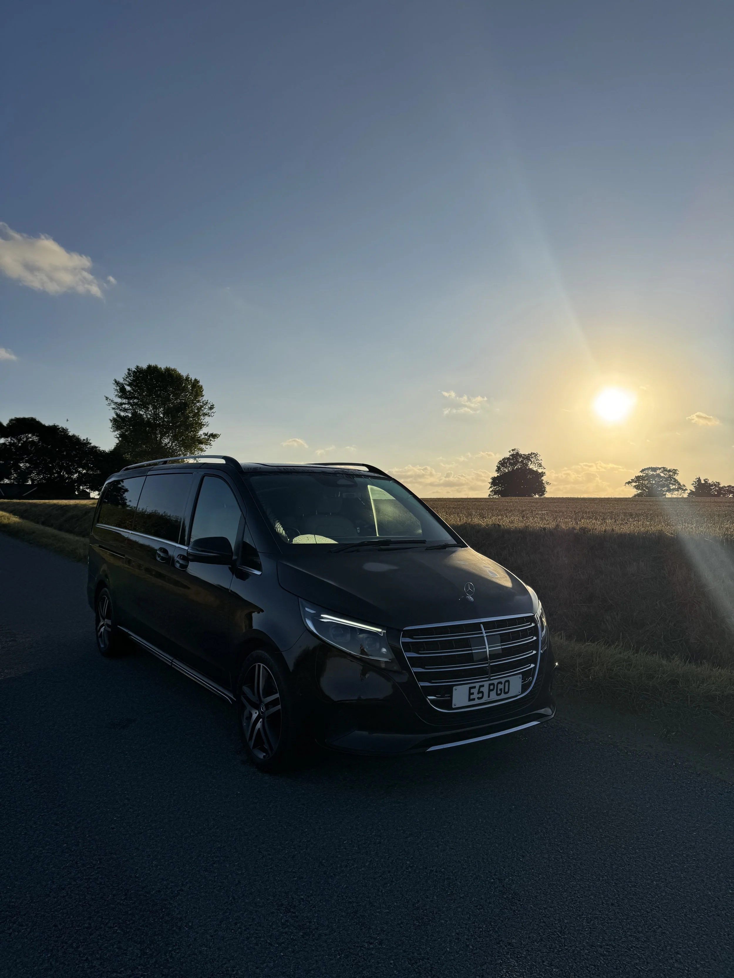 Norfolk Chauffeur Services Travel - Black Mercedes-Benz parked on a narrow country road with a large tree and green fields in the background.