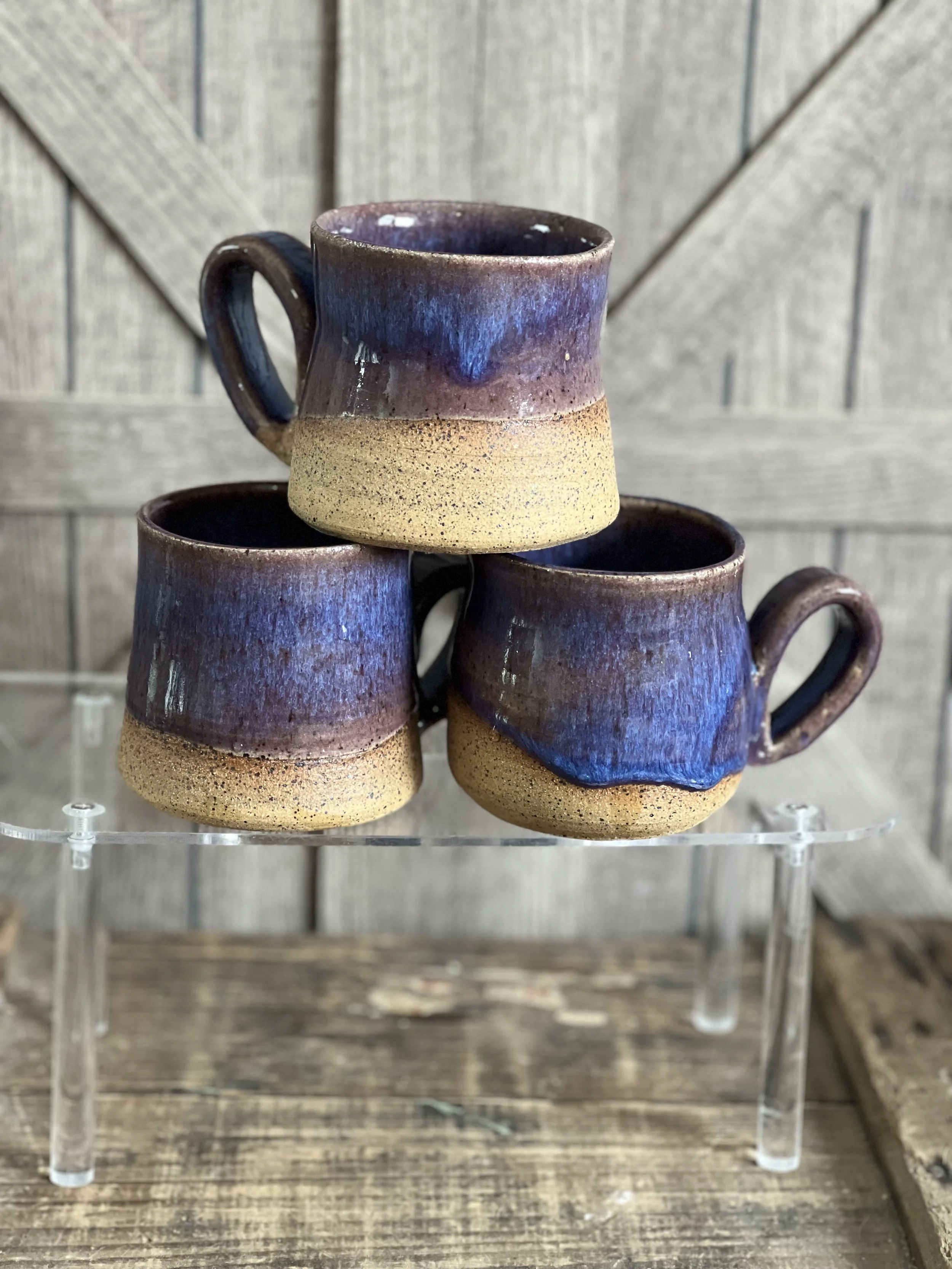 Three handmade ceramic cups with a purple and beige glaze, stacked on a clear acrylic stand against a wooden background.